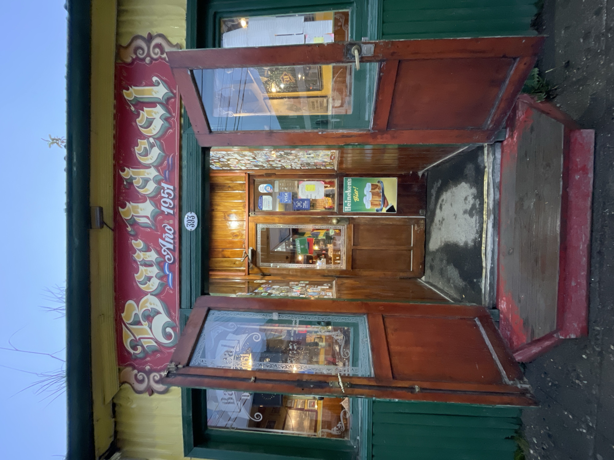 The image shows the open double doors of a business in Ushuaia, Argentina. The building is painted in yellow and green with a red vertical banner on the left featuring ornate lettering and the text "Año 1951" (Year 1951). A small white oval sign with the number "393" is visible on the building's exterior.
The doors are made of dark wood with glass panels, decorated with etched designs. Inside the doorway, there are signs for VISA and other credit cards. A Heineken beer advertisement is also visible. A wooden door is at the back of the entryway. The entrance has a raised wooden step, painted partially in red. The surrounding environment is a paved area with a view to the sky and bare tree branches.