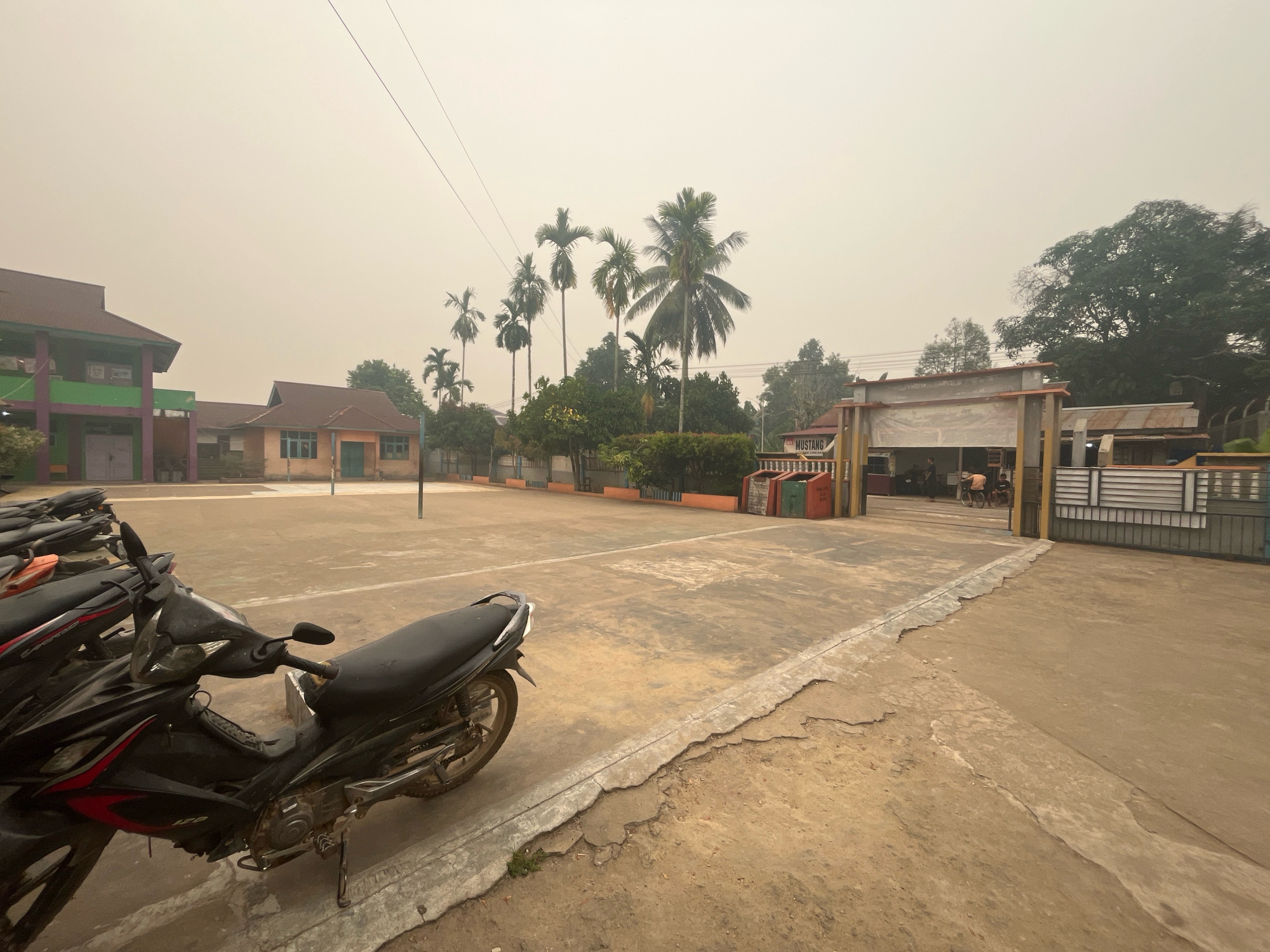 The image depicts an outdoor scene, likely a schoolyard or public space, in Meliau Hulu, Indonesia. The sky is hazy with an unnatural, pale yellow hue, suggesting an environmental concern such as smoke or smog.
In the foreground, several parked motorcycles are visible, with a prominent black and red motorcycle on the left side. The ground is a mixture of cracked concrete and dirt.
In the middle ground, a paved area, possibly a sports court, is marked with lines. A volleyball net stands on the court. Beyond the court, several buildings are visible, appearing to be low-rise school or administrative structures with red tiled roofs and some with green and purple exterior paint. Palm trees and other foliage are scattered throughout the area.
To the right, an entrance gate with a "MUSTANG" sign is present, leading into a building or complex. The gate itself is a combination of metal bars and solid panels. A few individuals are faintly visible within the entrance area. The overall atmosphere is muted due to the hazy sky.