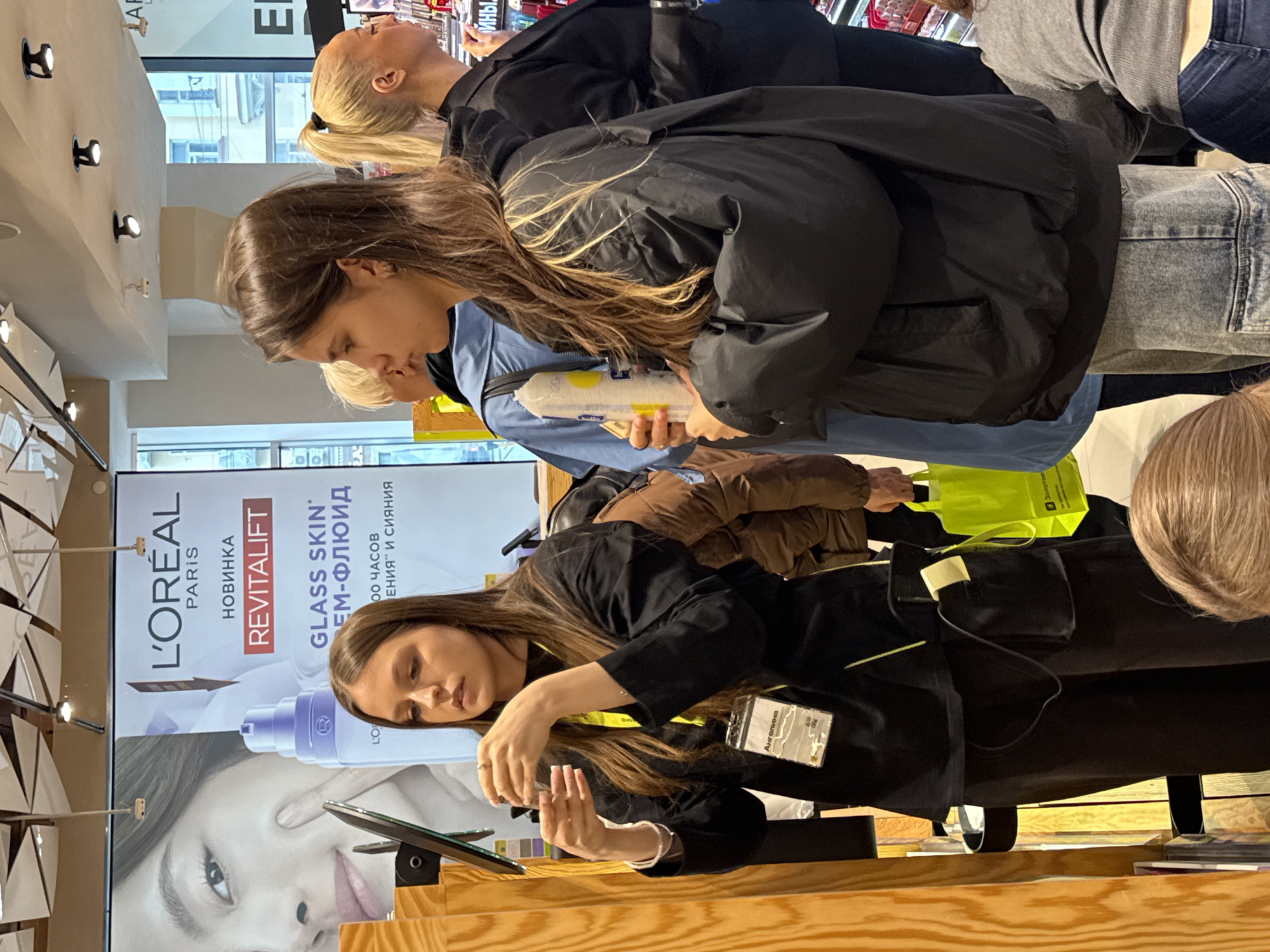 The image, taken in Kazan, Russia, shows three women in a store. One woman, wearing a black jacket, is holding a white bottle with yellow accents and is looking down. Another woman, with blonde hair pulled back in a ponytail, is looking up. The third woman is looking at a digital device. A L'Oréal Revitalift advertisement is in the background, featuring a woman's face and product information in Russian. A name tag labeled "Ангелина" is visible on one of the women's jacket. A green tote bag and various clothing items are also visible.