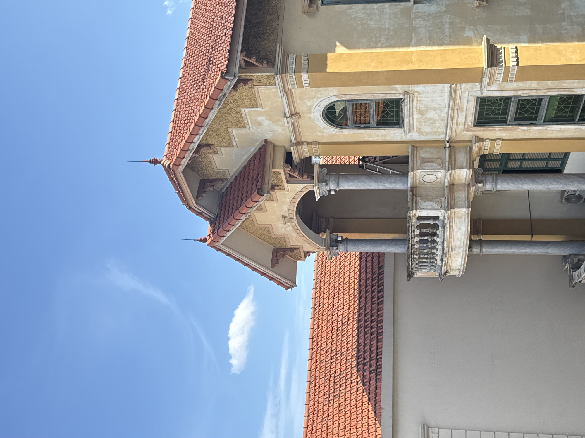 The image shows a partial view of a building with distinct architectural features. The building has a red-tiled roof, yellow-painted walls, and a colonnaded balcony with ornate carvings. The design suggests European influence. The sky above is blue with wispy clouds. Without further context, it is not possible to confirm that the location is Hà Nội, Vietnam.