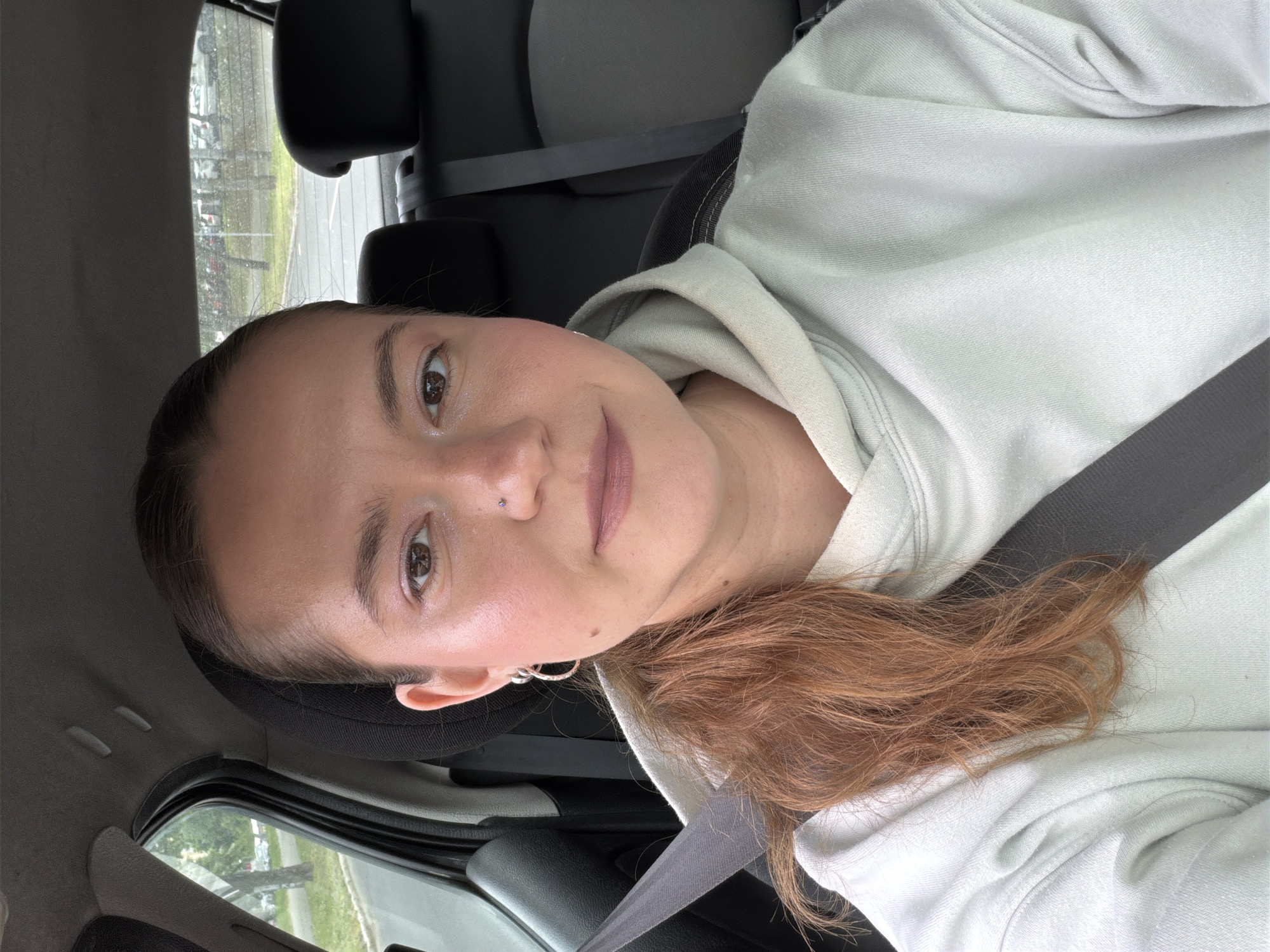 A close-up selfie shows a young woman seated inside a car, looking directly at the camera with a neutral and slightly pleasant expression. Her light brown to auburn hair is styled in a pulled-back ponytail, with some loose strands resting on her shoulder and chest. She is wearing a light beige or off-white hooded sweatshirt and a dark grey seatbelt diagonally across her body. Distinctive features include a small silver hoop earring in her left ear and a tiny silver stud nose piercing.
The interior of the car is visible, with dark seats and headrests behind her. Through the car windows, an outdoor scene unfolds, revealing green trees, a road or parking area, and hints of buildings in the distance, consistent with an urban or suburban environment like the Vozdovac Urban Municipality in Serbia. The lighting suggests it is daytime, possibly an overcast day, and some surfaces outside the car appear damp, indicating recent rain or cloudy weather. The image is oriented horizontally, as if the camera was rotated during capture. There is no visible text in the media file.