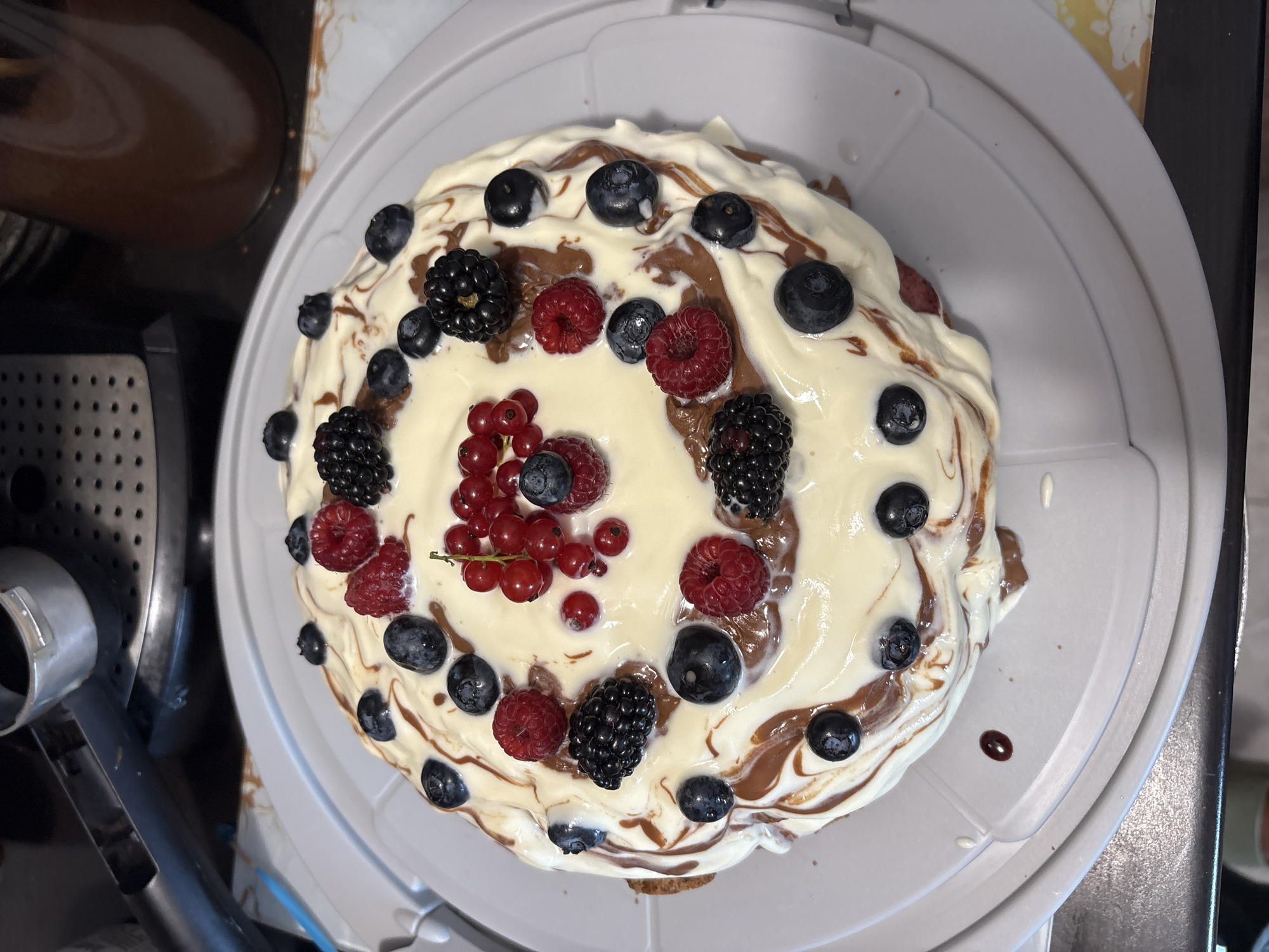 An overhead shot shows a cake with white frosting and chocolate drizzled on top, decorated with an assortment of berries including raspberries, blackberries, blueberries, and red currants. The cake is on a light gray cake stand. In the blurred background, a coffee machine with a portafilter can be seen, along with a dark brown bottle. The overall scene appears to be indoors, likely in a kitchen environment, but no specific city or location can be identified.
