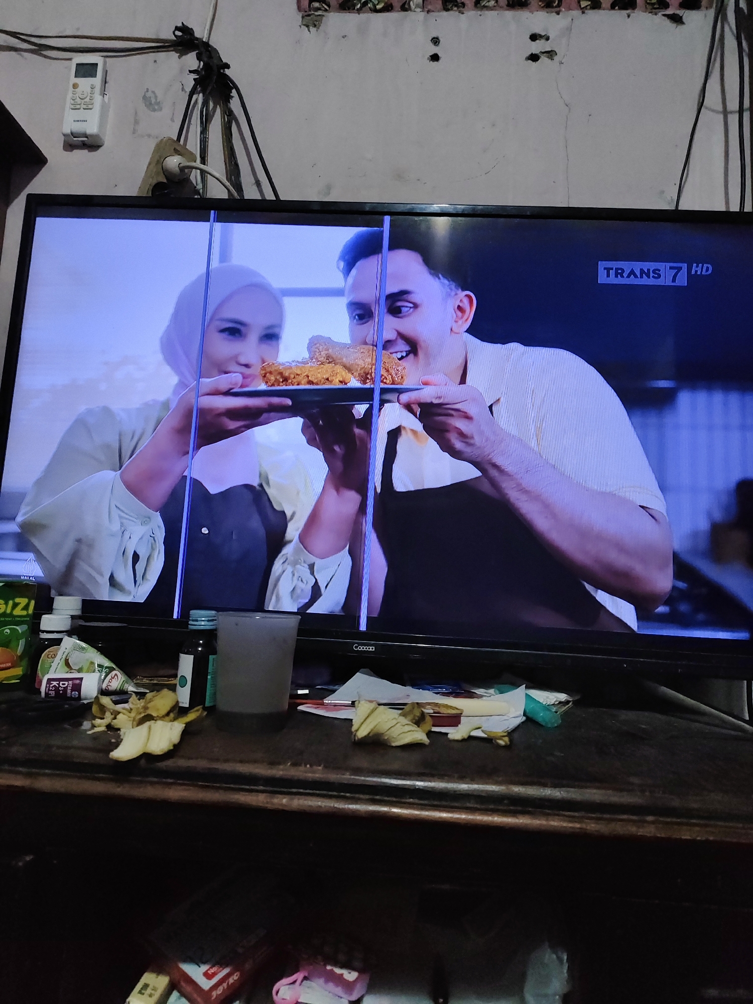 The image shows a close-up of a television screen displaying a scene from what appears to be a cooking show or advertisement. On the screen, a woman wearing a light-colored hijab and a man in a light-colored, textured shirt are holding a plate of fried chicken. The woman is on the left, and the man is on the right, smiling broadly. The television is placed on a dark wooden surface, cluttered with various items. Among the items are bottles of supplements, what looks like a tube of toothpaste, and some discarded banana peels. The background of the image shows a wall with exposed wires and an electrical outlet, suggesting a somewhat disorganized or lived-in environment, possibly a residence in South Jakarta, Indonesia. The overall scene is dimly lit, with the focus on the television screen.