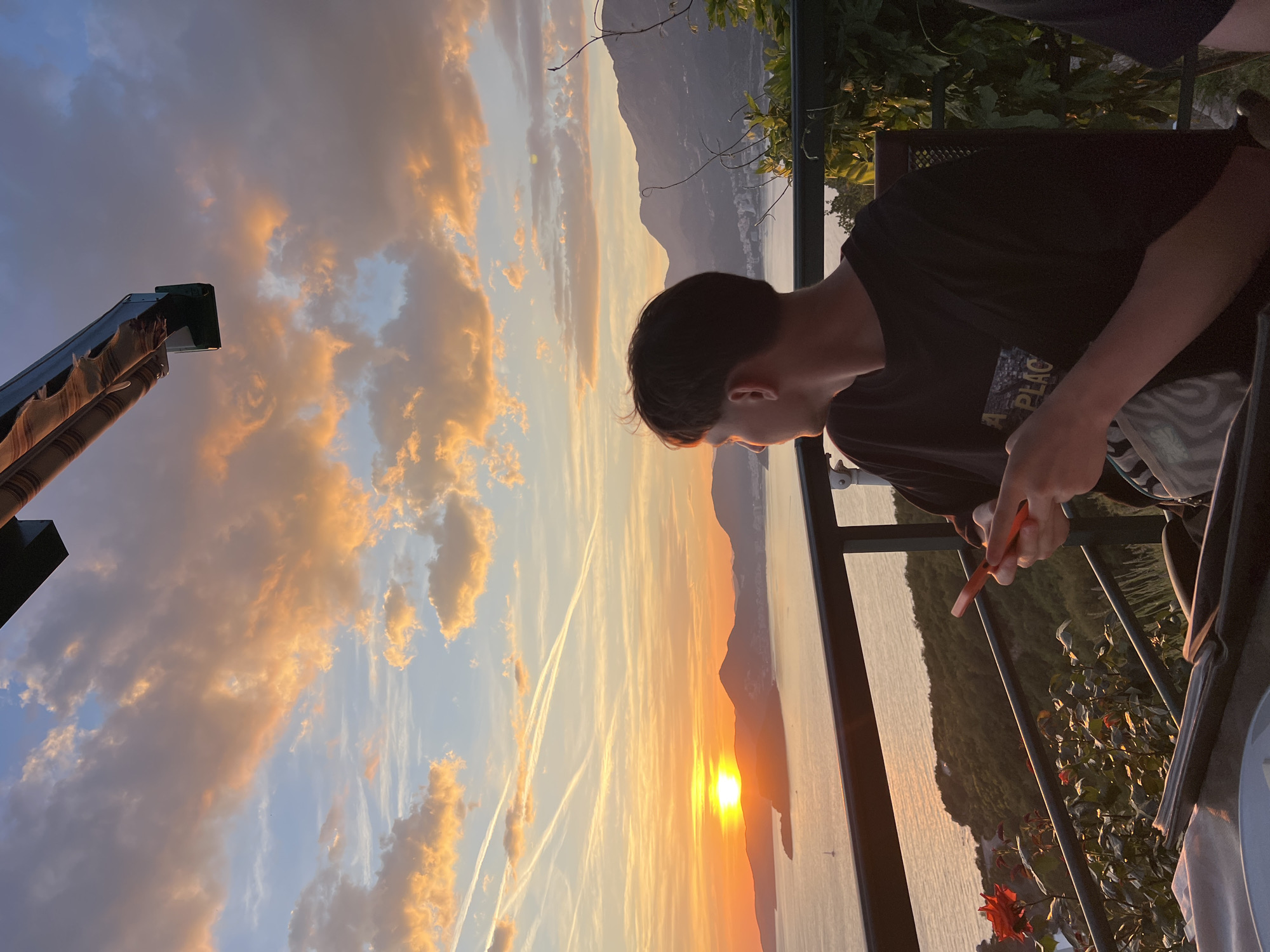 At dusk in Blizikuće, Montenegro, a person wearing a dark t-shirt holds an orange lighter, looking toward a sunset over water. The view includes mountains and clouds illuminated by the setting sun, and is framed by a balcony with greenery. An awning is partially visible in the upper left corner.