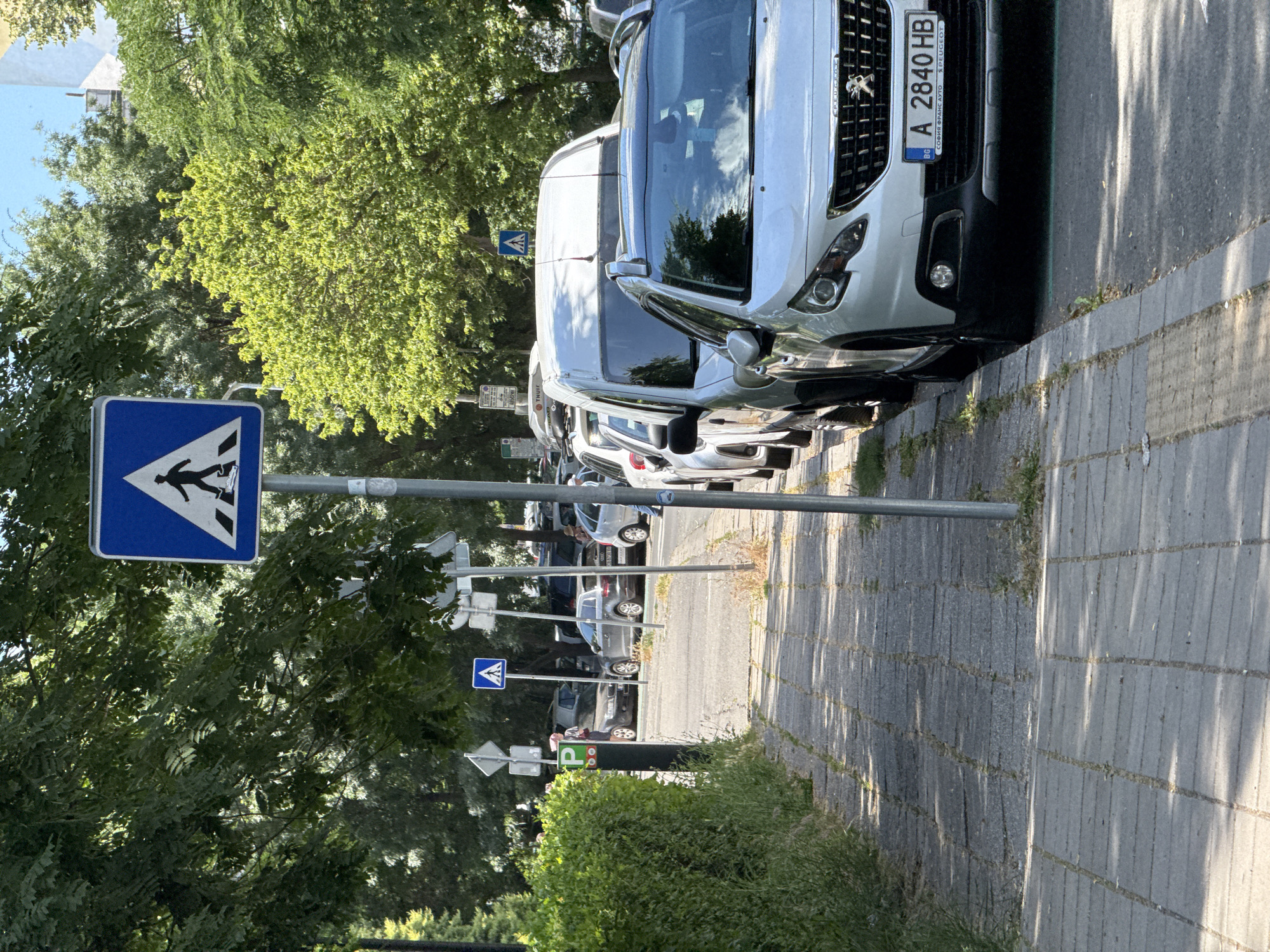 The image depicts a street scene in Burgas, Bulgaria, likely during the daytime on a clear, sunny day, as indicated by the strong shadows cast on the paved walkway. The focus is on a row of parked cars along a street lined with lush green trees. A prominent pedestrian crossing sign, a blue square with a white silhouette of a person walking, is visible on the left side, suggesting this is a designated crossing area. Further down the street, another pedestrian crossing sign and a parking sign with a green "P" are visible, indicating parking regulations are in effect.
The cars are parked parallel to the sidewalk. The closest vehicle, a silver Peugeot with a Bulgarian license plate "A 2840 HB", is clearly visible, with text on the license plate including "BG" and "PEUGEOT". The street is paved with cobblestones, and a strip of grass runs along the edge of the walkway. The trees provide ample shade, creating a dappled lighting effect on the ground. The scene appears peaceful and ordinary, with no people actively engaged in any notable activity. The overall impression is a typical urban streetscape during a pleasant day.