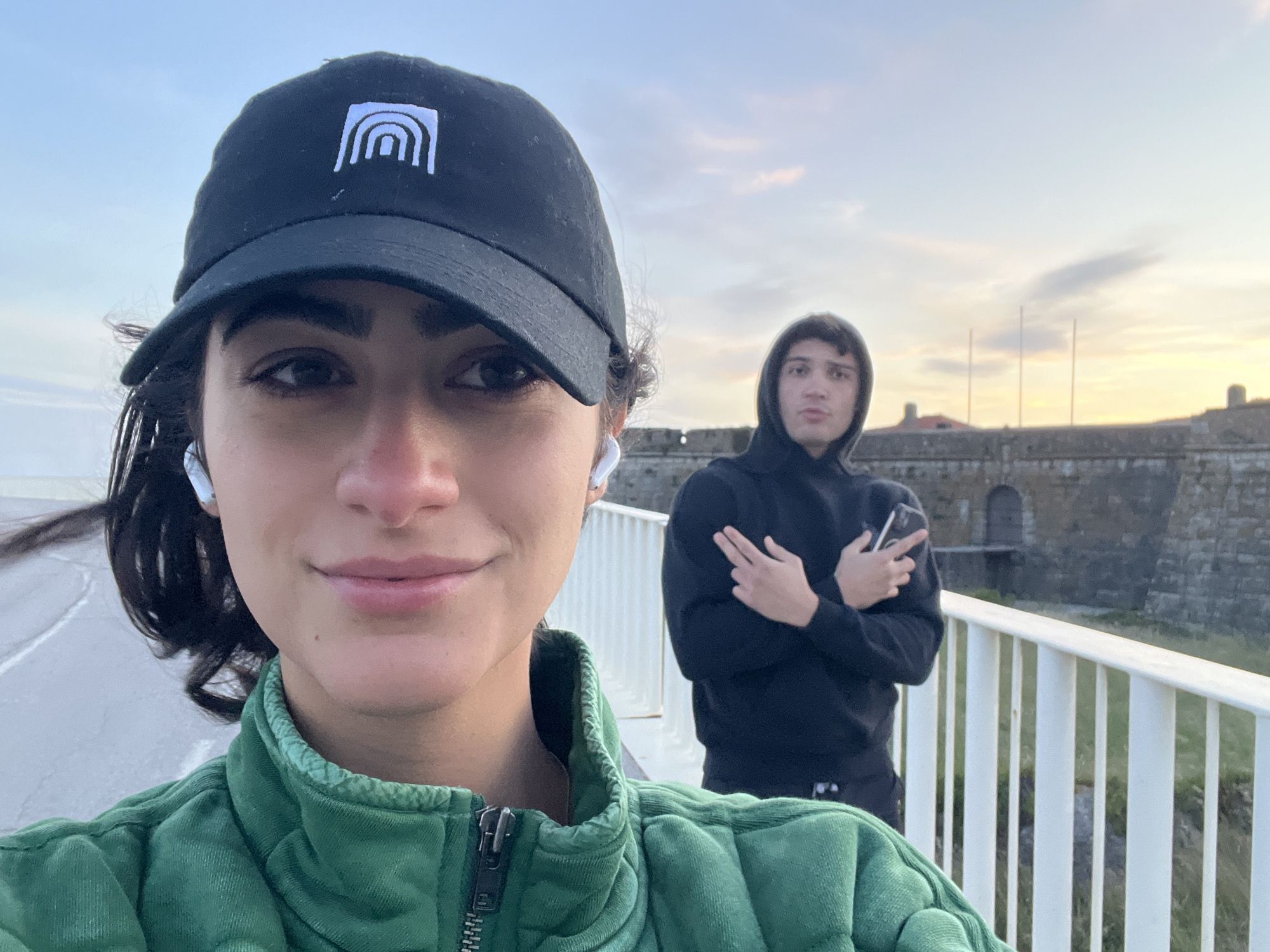 A woman wearing a black baseball cap with a white logo, a green jacket, and white earbuds takes a selfie outdoors. In the background stands a young man in a black hoodie making a gesture with his fingers. They are standing on a white bridge with a stone building behind them. The image appears to be taken near sunset with the sky showing hues of orange. The location is the Fortaleza de São João da Foz in Porto, Portugal.