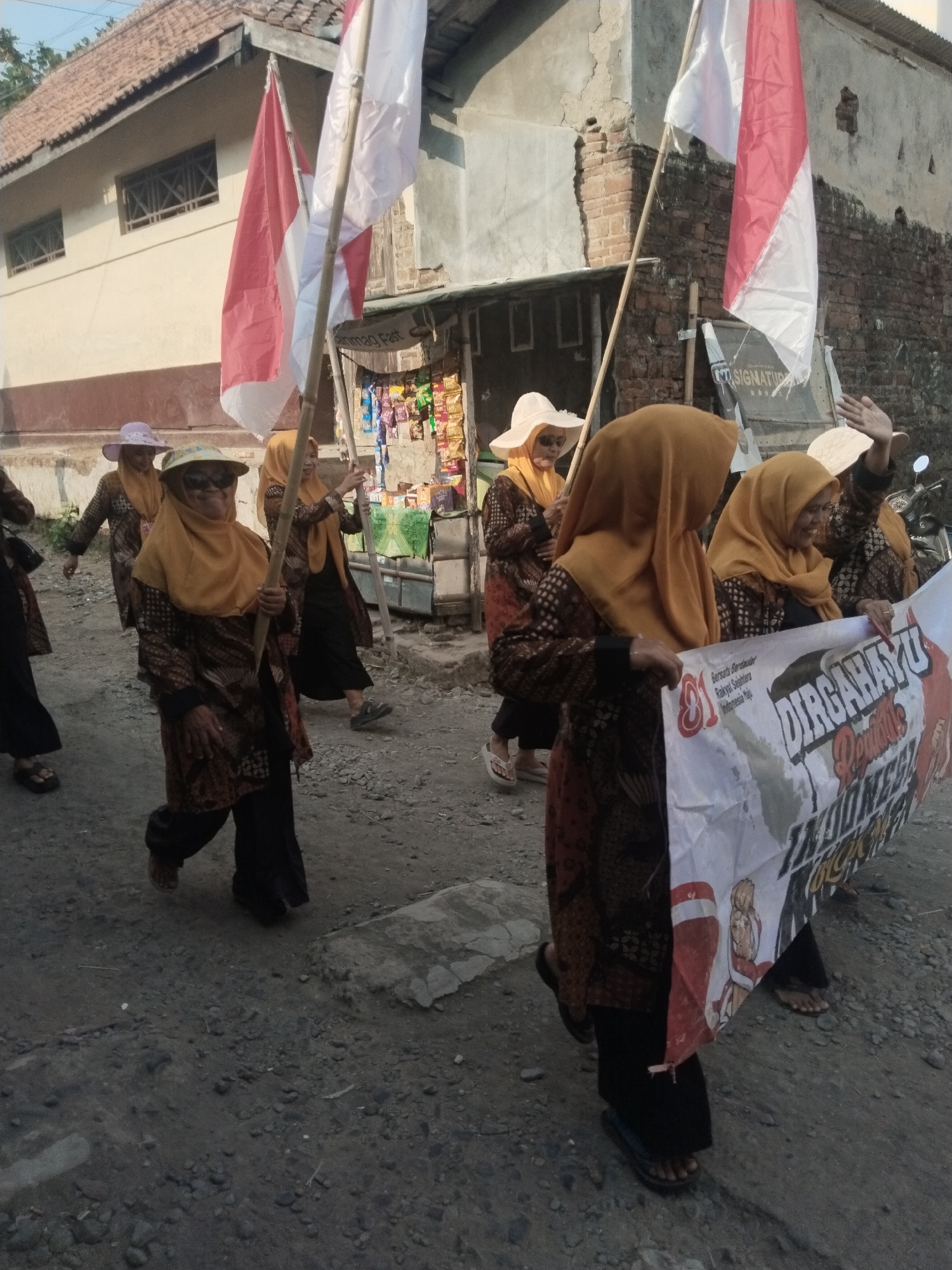 A group of women are walking in a procession down a dirt road in Jembayat, Indonesia, holding Indonesian flags and a banner. The woman in the foreground is wearing a yellow hijab and a patterned brown and orange batik tunic, and she is holding a wooden pole. Behind her, other women are also wearing hijabs and batik clothing. They are carrying two Indonesian flags, red and white, and a banner that reads "DIRGAHAYU REPUBLIK INDONESIA". The road is unpaved and has gravel and dirt. Buildings with tiled roofs are visible in the background. The sky is clear.