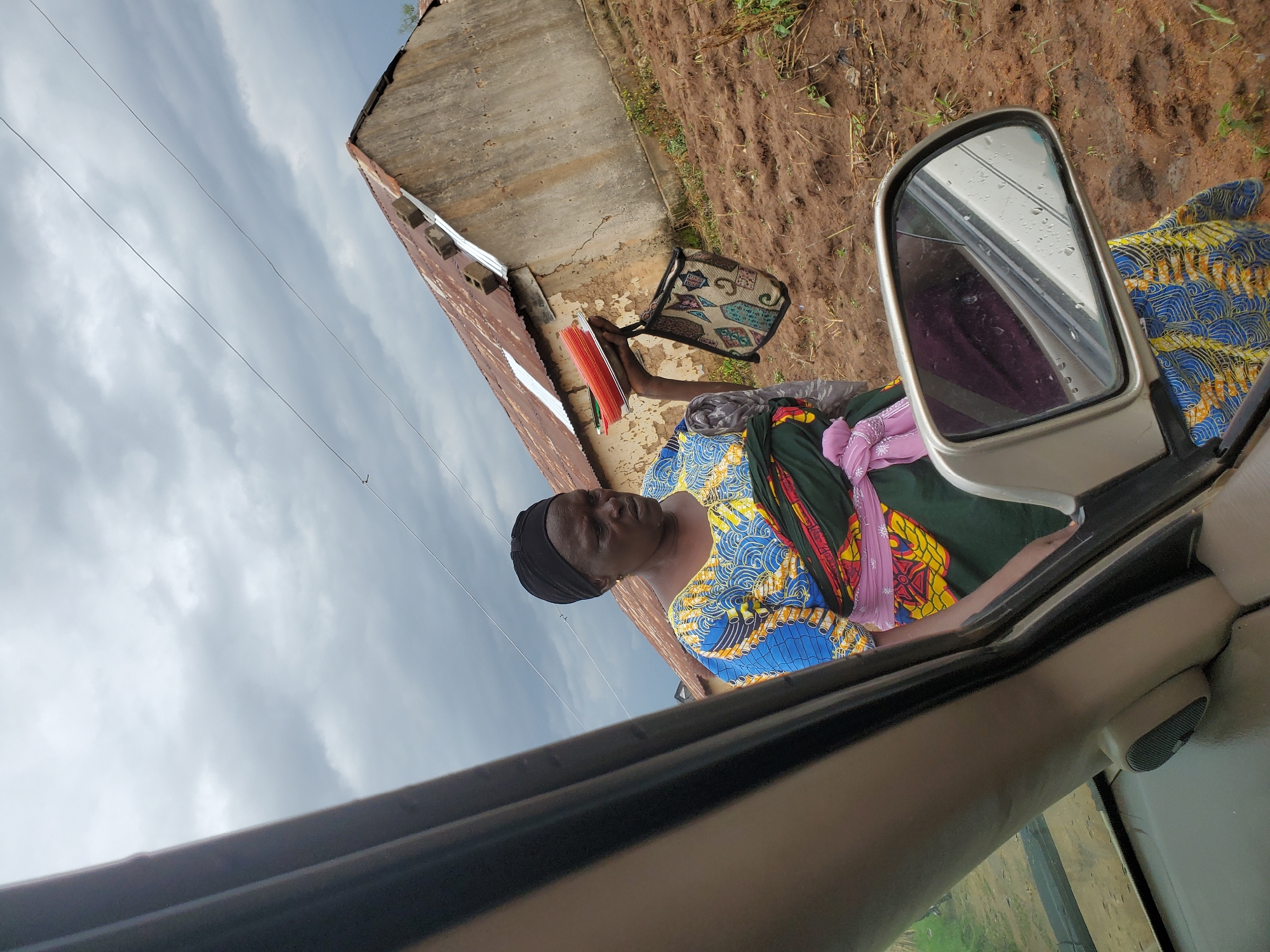 An overhead, medium shot captures an older Black woman dressed in a vibrant, patterned traditional outfit and a black headscarf. She is standing in front of a weathered, dilapidated building with a rusty corrugated metal roof. The ground is muddy and uneven, indicative of recent rainfall. The woman is holding a stack of orange items, possibly books or folders, and a small, patterned bag. Her gaze is directed downwards. In the foreground, the side mirror and part of the interior of a vehicle are visible, suggesting the photo was taken from inside a car. The sky is overcast with heavy, grey clouds. The scene appears to be in a rural or semi-urban area, consistent with parts of Jos, Nigeria.