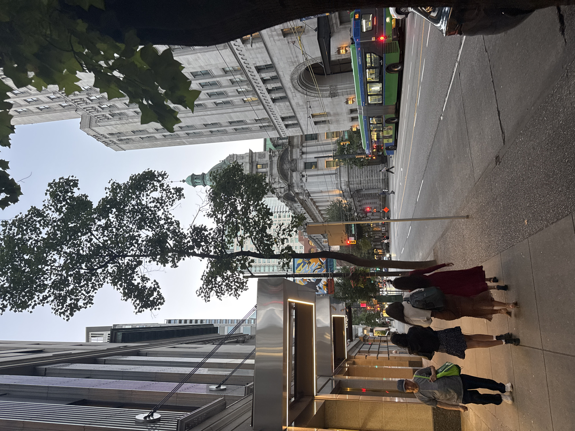 This image depicts a street scene in downtown Vancouver, Canada. The view is from a sidewalk looking east down a city street, with tall buildings on either side. On the left, there is a modern building with a metallic facade and illuminated overhangs. Across the street, on the right, a grand, older stone building with arched entrances and a domed roof is visible, alongside a taller, more contemporary stone building. A green and blue public transit bus is moving along the street. Several people are walking on the sidewalk in the foreground, including a man in a grey shirt and blue pants, and three women walking together. The sky is overcast, suggesting it is either early morning or late afternoon. The overall impression is a busy urban environment.