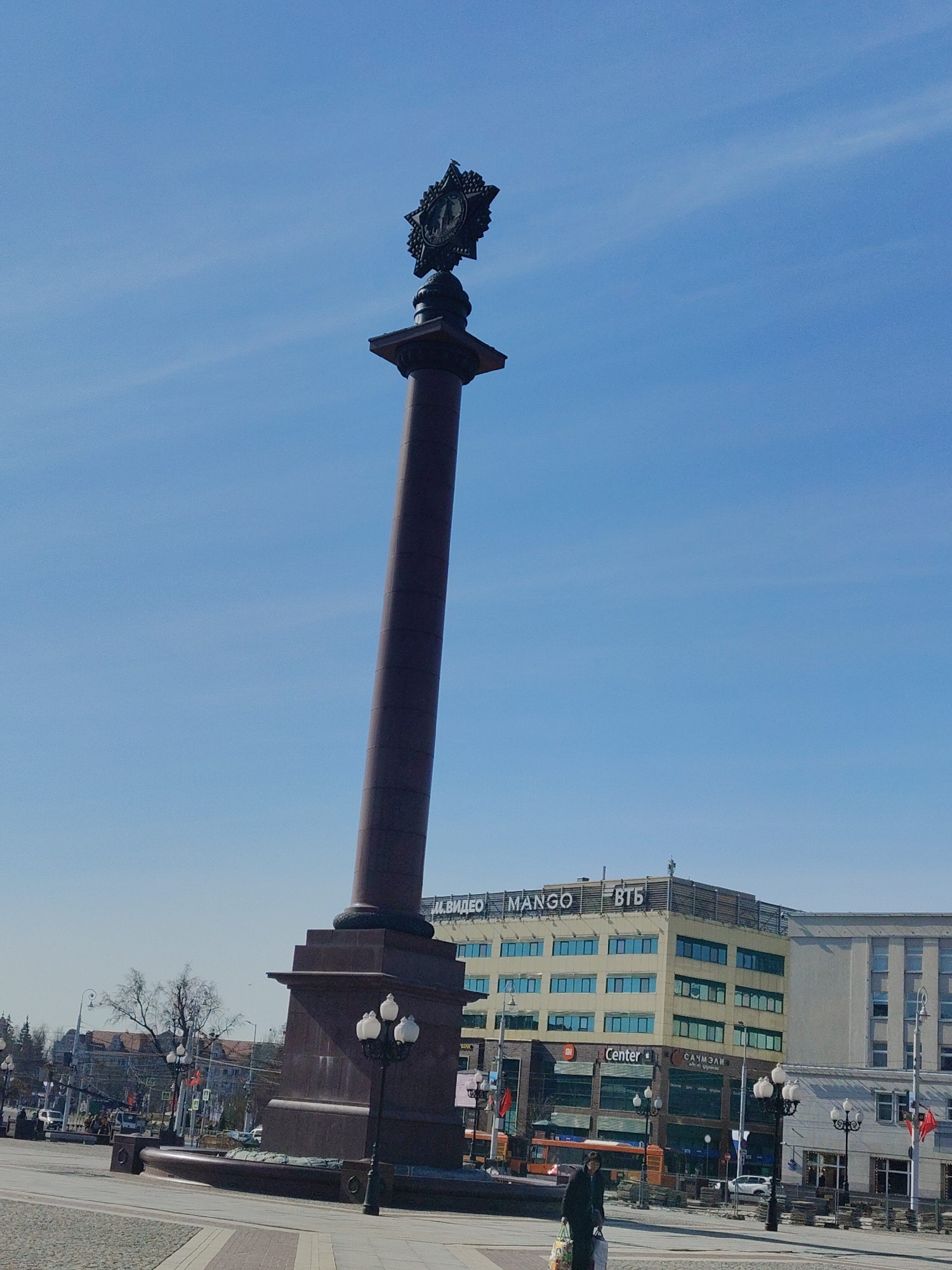 The image captures a tall, dark-colored monument in an urban environment. The monument, with a column topped by a star-like emblem, dominates the frame against a clear blue sky with light clouds. At its base, there are decorative elements, including a water feature. Street lamps are positioned around the monument.
In the background, a multi-story building is visible with signs in Cyrillic script, indicating commercial presence. Another building in a different style is next to it.
A few individuals and vehicles can be seen in the lower part of the image. The paving of the square is visible in the foreground. The overall scene suggests a public square in a city with Soviet-era influenced architecture.
Location: Unknown, Unknown.