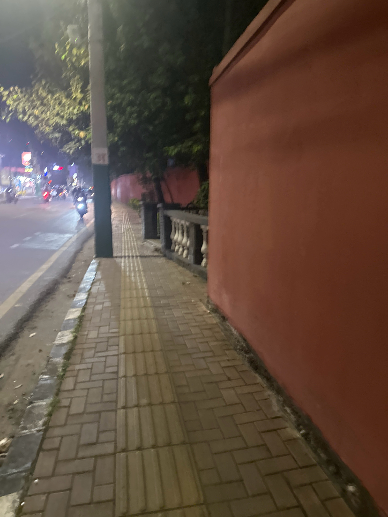 This is a low-light, slightly blurred image of a sidewalk at night in Lalitpur, Nepal. A reddish-orange wall runs along the right side of the frame, separating the sidewalk from an unseen area. The sidewalk is paved with rectangular bricks laid in a herringbone pattern. There are some indications of a street to the left, with vehicles, possibly motorcycles, visible in the distance, their lights creating streaks of color. A lamppost is present on the sidewalk near the center of the image. A low decorative railing with balusters is visible in the background along the wall. The overall impression is a street scene in an urban environment at night.