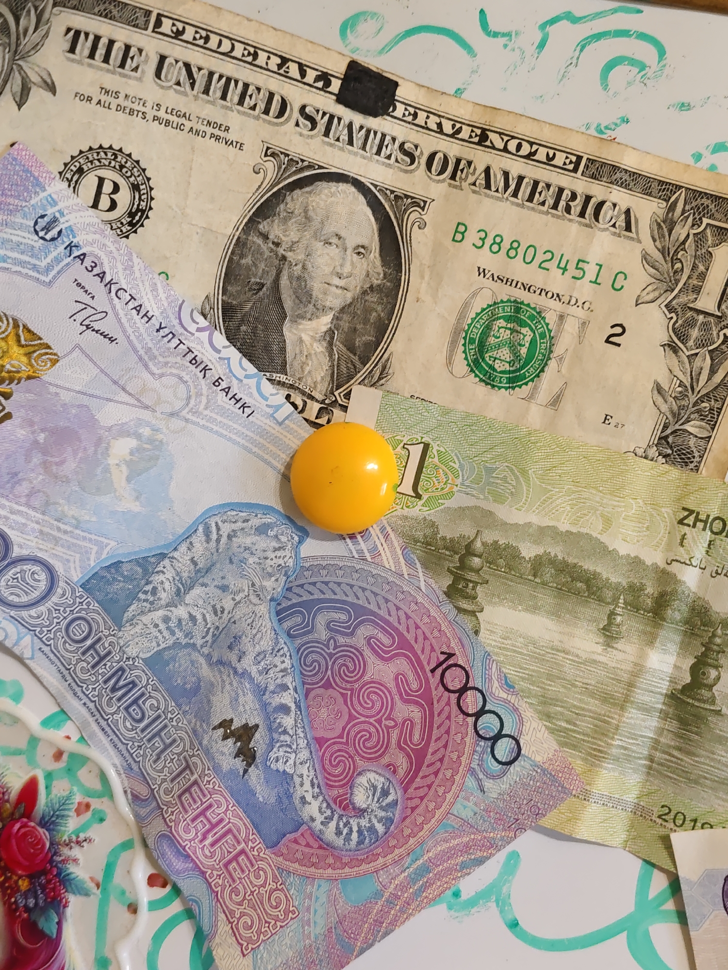 The image shows a close-up of several banknotes, including a US dollar bill and what appear to be Kazakhstani tenge and Chinese yuan. The banknotes are overlaid on a white surface with turquoise abstract designs. A small yellow sphere is also visible. The image is taken from a slightly elevated angle, and the lighting suggests an indoor setting. The banknotes are somewhat creased and worn. The US dollar bill is partially visible, featuring a portrait of George Washington and text indicating it is legal tender. The Kazakhstani tenge banknote has intricate designs, including what appears to be a snow leopard. The Chinese yuan banknote displays a landscape with pagodas and a body of water. There are no people or animals visible in the image, and no specific landmarks of Rostov-on-Don, Russia, are identifiable.