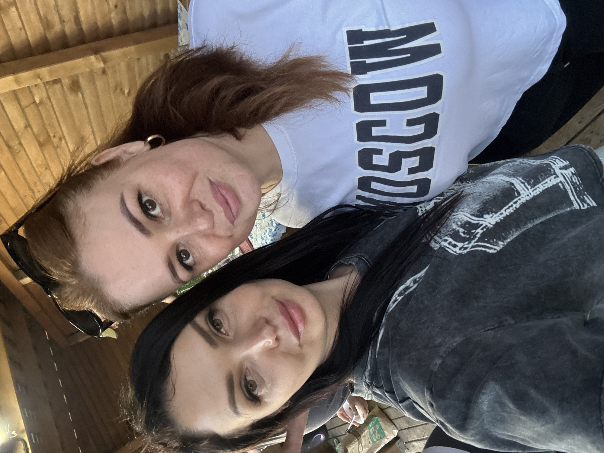 Two women are taking a selfie outdoors under a wooden structure. The woman on the left has long, dark hair and is wearing a dark, distressed t-shirt with white graphics. The woman on the right has reddish-brown hair pulled back and is wearing a white t-shirt with the word "MOSCOW" in large, black, block letters. She has sunglasses perched on top of her head. The background shows wooden beams and planks of the structure, suggesting it is a covered patio or gazebo. To the left of the women, a brown paper bag and some disposable cups are visible on a wooden surface, indicating a casual outdoor setting. The lighting suggests it is daytime. This scene could be located anywhere in Votkinsk, Russia, or elsewhere, as there are no distinguishing landmarks.