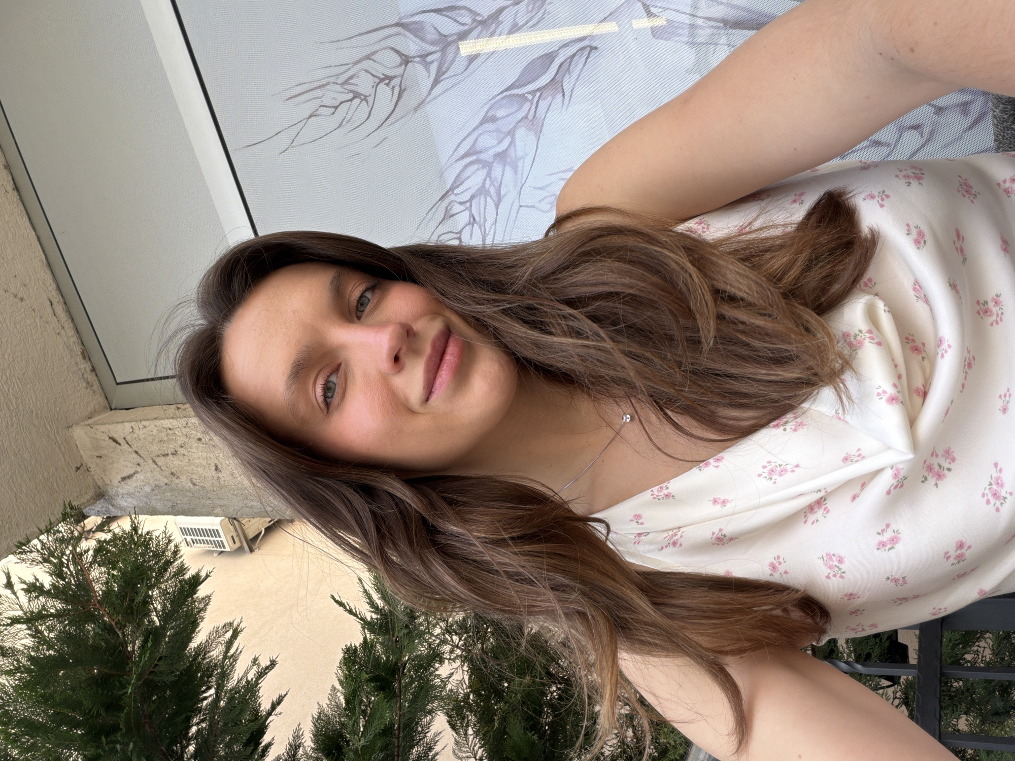 The image is a selfie of a young woman taken outdoors in Budva, Montenegro. She is the primary subject, looking directly at the camera with a soft, natural smile. She has long, wavy brown hair, light-colored eyes, and appears to be wearing minimal makeup. Her head and upper torso are visible.
She is dressed in a light cream or off-white top featuring a delicate, scattered pattern of small pink floral prints. A thin silver necklace with a tiny, round pendant is visible around her neck.
The setting appears to be a balcony or an area adjacent to a building. In the background, to her left, are several vibrant green coniferous trees. Above the trees and behind her head, a light-colored building structure is visible, with an external air conditioning unit mounted on a wall. To her right, a large window or glass panel is present, featuring a subtle, light grey etched or printed design resembling organic forms like branches or leaves.
The time of day appears to be daytime, characterized by bright, natural illumination, suggesting clear weather. The woman's expression conveys a calm and contented emotion. There is no visible text in the image.