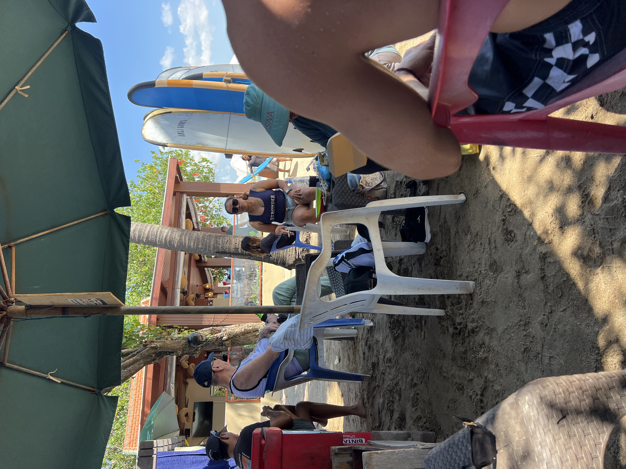 In Kuta, Indonesia, an outdoor beach cafe scene unfolds under a large, green umbrella. Several people are seated on chairs on the sandy ground. In the foreground, the arm and shoulder of an individual wearing a checkered swim trunk are visible. To the left, a person in a black t-shirt and shorts sits near a red cooler. A man in a white tank top and blue shorts, wearing a blue baseball cap, is seated in a blue plastic chair. Another person in a blue tank top and denim shorts is seated across from him. Two surfboards, one blue and one white, stand upright behind the seated individuals. In the background, a building with an orange roof and wooden pillars appears to be part of the cafe establishment, with other patrons visible inside. The sky above is blue with scattered clouds.