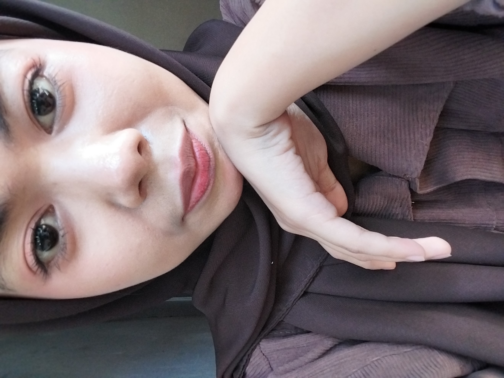 A close-up, frontal portrait captures a young person wearing a dark brown hijab and a brown textured shirt. Their face is turned slightly upwards, and their chin rests on their left hand, which is positioned near their mouth. The individual's eyes are open and looking towards the camera, with visible eyelashes and subtle eye makeup. The lips are slightly parted, revealing a pinkish hue. The background consists of a neutral, out-of-focus wall, suggesting an indoor setting. The lighting appears to be soft, illuminating the subject's face and highlighting the texture of their clothing and hijab. This image was taken on a mobile phone in Tibojong, Indonesia.
