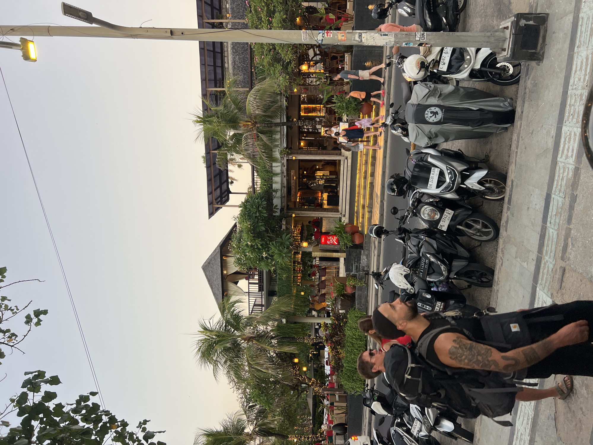 A wide shot captures a street scene in Kuta, Indonesia, with a focus on a row of parked motorcycles in the foreground and a multi-story building with an outdoor dining area behind them. The building appears to be a restaurant or bar, with tropical plants and string lights decorating the facade. Several people are visible on the sidewalk, some walking and others seated at tables. The sky is a pale orange, suggesting either dawn or dusk. The motorcycles are a mix of models and colors, with some covered by protective tarps. The street itself is paved, and a sidewalk runs alongside the parked vehicles. The overall atmosphere is one of a bustling tourist destination.
