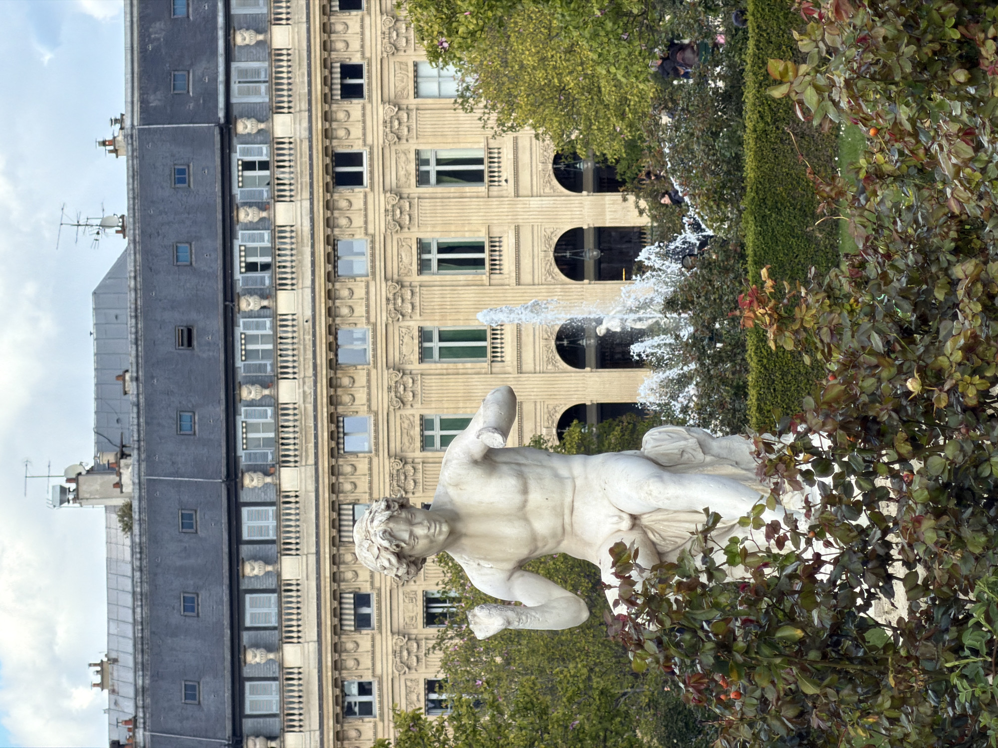 The image presents an outdoor scene in the Jardin des Tuileries, Paris, France, with a prominent white marble statue in the foreground. This statue depicts a youthful, nude male figure seated on a draped base, with curly hair, an athletic build, and visible genitalia. His left arm is bent with a clenched fist, while his right forearm is missing. The statue appears weathered and faces slightly right. It is partially obscured by dense rose bushes featuring dark green and reddish leaves with orange rosehips.
Behind the statue and foliage, a fountain is active in the midground, spraying white water jets into the air. Further back, occupying the entire background, is a grand, multi-story building exhibiting classical Parisian Haussmannian architecture. Its facade is a light ochre or beige stone, adorned with fluted pilasters, cornices, and decorative friezes. The lower levels feature large arched windows and entrances, while upper floors display rows of rectangular windows, some with light blue shutters and small balustrades. The building is topped with a dark grey slate mansard roof, punctuated by numerous dormer windows, chimneys, and various antennae.
Scattered within the midground, behind the hedges and near the fountain, several indistinct individuals are visible, either standing or walking. The sky above is overcast, a mix of light grey and white clouds.