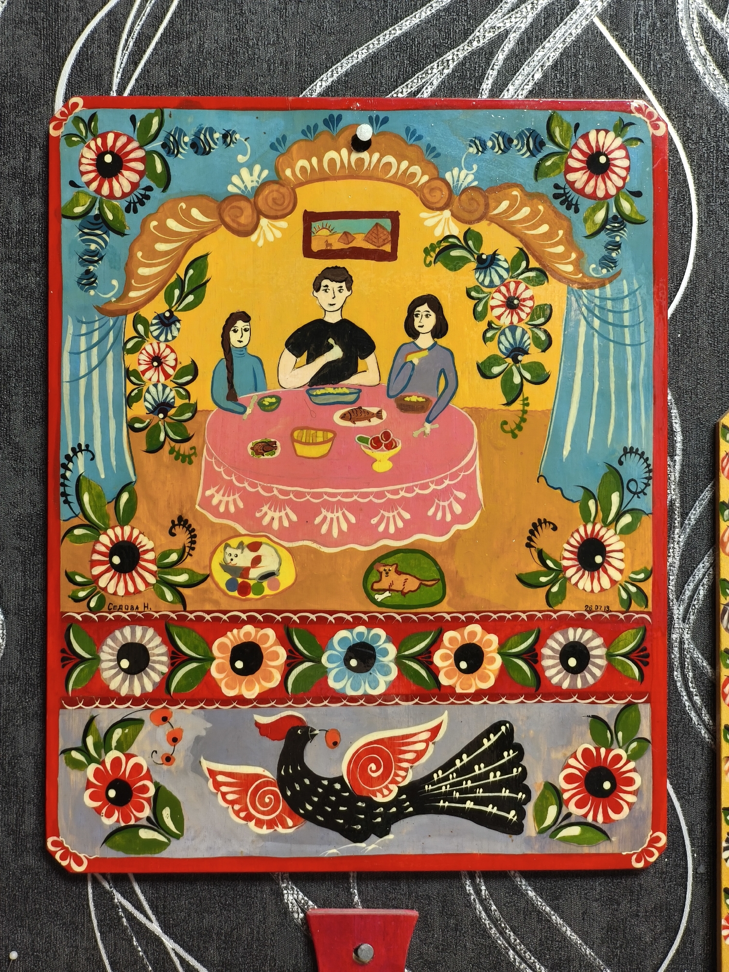 This is a vibrant folk art painting, likely on a wooden plaque, characterized by its bold colors and stylized forms. The central scene depicts three individuals – a man and two women – gathered around a pink, lace-edged table, appearing to share a meal. The man, wearing a black shirt, is seated in the middle, while a woman in a blue turtleneck sits to his left, and another woman in a gray top to his right. Dishes on the table include what looks like a whole fish, bowls of food, and a fruit bowl.
The scene is set indoors, against a bright yellow wall. Above the table, an ornate, arch-shaped canopy in shades of orange and brown frames the space, adorned with flowers and swirling motifs. Blue curtains with vertical stripes flank the scene. A small framed picture of pyramids is visible on the back wall, suggesting an Egyptian landscape. Two domestic animals are featured in circular beds on the floor: a white cat with red markings on the left and a brown dog on the right.
The entire composition is richly decorated with numerous stylized flowers and foliage in various colors (red, white, blue, orange, green) that frame the main scene and form a prominent border at the bottom. The bottom-most section of the painting features a large, stylized black bird with red and white wings, holding a red berry in its beak, set against a light gray background, also surrounded by flowers.
The painting's style is reminiscent of Eastern European or Slavic folk art, often seen in decorative painting. The colors are striking and harmonious, creating a cheerful atmosphere.
Visible text includes what appears to be the artist's name, "СЕДОВА Н." (SEDOWA N. or SEDOVA N.), written in Cyrillic in the bottom-left floral border section. A date, "26.07.19" (July 26, 2019), is inscribed in the bottom-right floral border area.