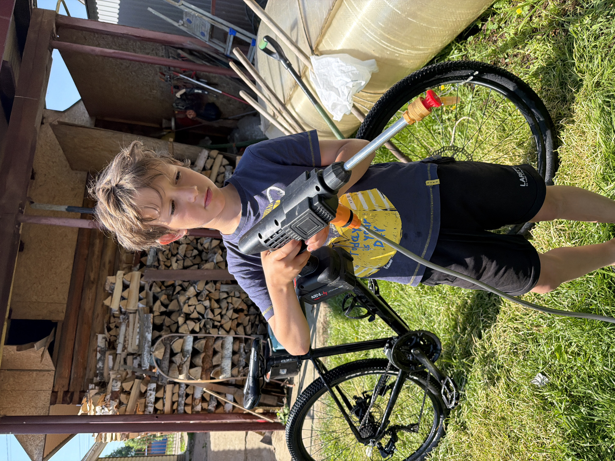 A young boy stands on grass in a rural area of Chaikovsky, Russia, cleaning a black bicycle with a cordless pressure washer. The background includes a woodpile, a ladder, and other yard implements leaning against a shed. The scene is sunlit, and the boy is looking directly at the camera.
