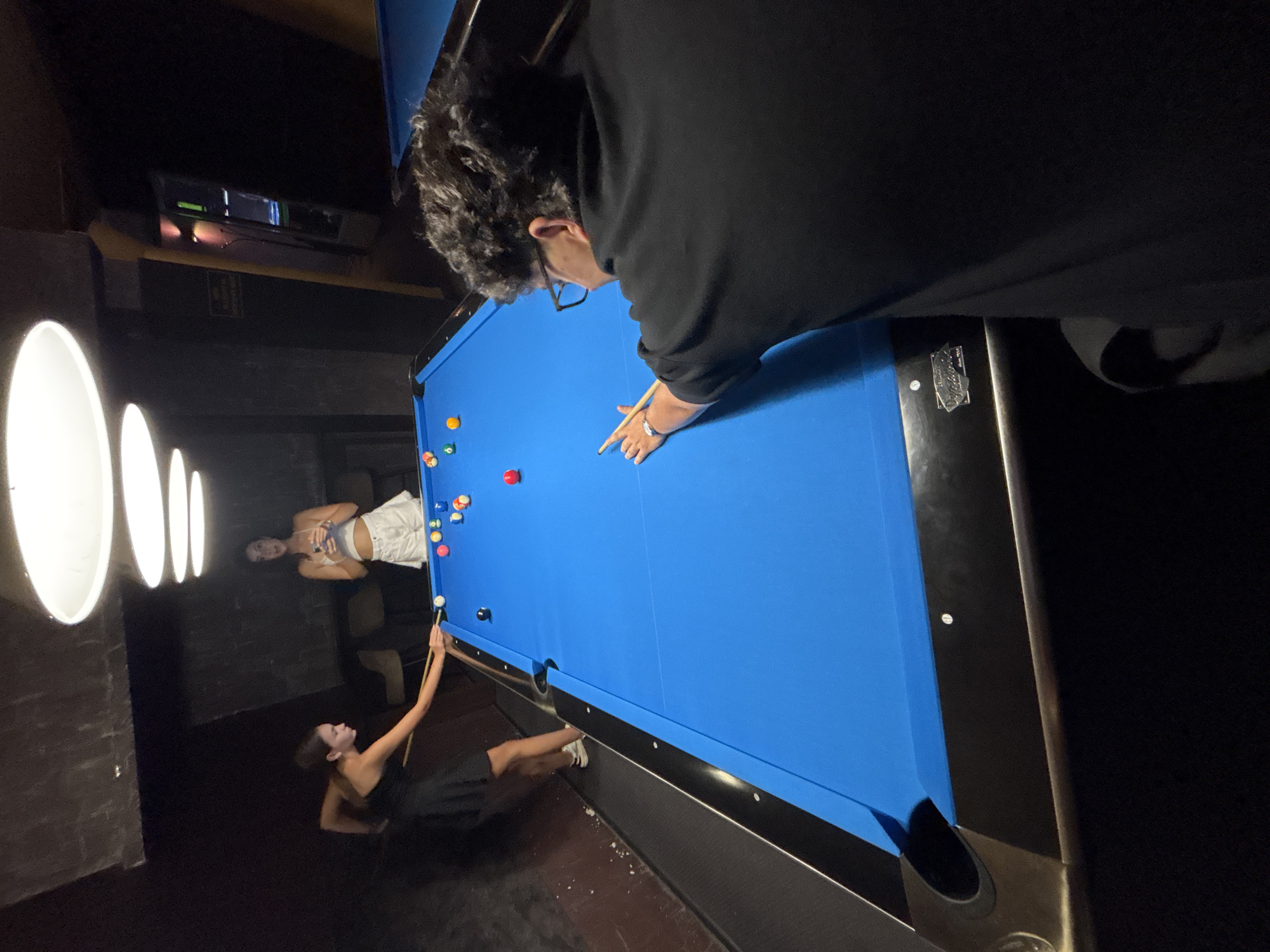 The image depicts a dimly lit indoor setting, possibly a bar or a game room, where a game of billiards is in progress. A bright blue billiard table dominates the foreground, with several billiard balls scattered across its surface. A man with curly hair and glasses is positioned on the right side of the table, leaning forward and aiming to strike a ball with a cue stick. On the left side of the table, a woman wearing a black dress is also engaged in the game, her cue stick extended towards a ball. In the background, another woman is visible, holding a drink and observing the game. The environment is characterized by dark walls, overhead lighting fixtures, and what appears to be a partial view of another billiard table on the far right. The scene is captured from a slightly elevated perspective, looking down at the billiard table and the players. The location is not specified in the image, and therefore cannot be determined to be in Vratsa, Bulgaria.