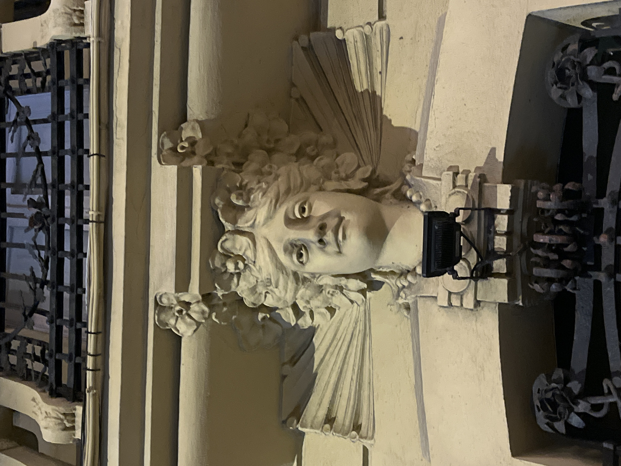 A close-up, angled shot depicts architectural detail, possibly from a building facade in A Coruña, Spain. A carved stone or plaster sculpture of a female head with floral and fan-like elements radiates outward from the face. A modern black light fixture is mounted near the sculpture. To the left, a window with a decorative metal grate is visible, while a darker, ornamental metalwork element exists on the right. The lighting appears to be artificial, casting shadows that highlight the relief of the carvings. The color palette is primarily muted beige, white, and dark gray.