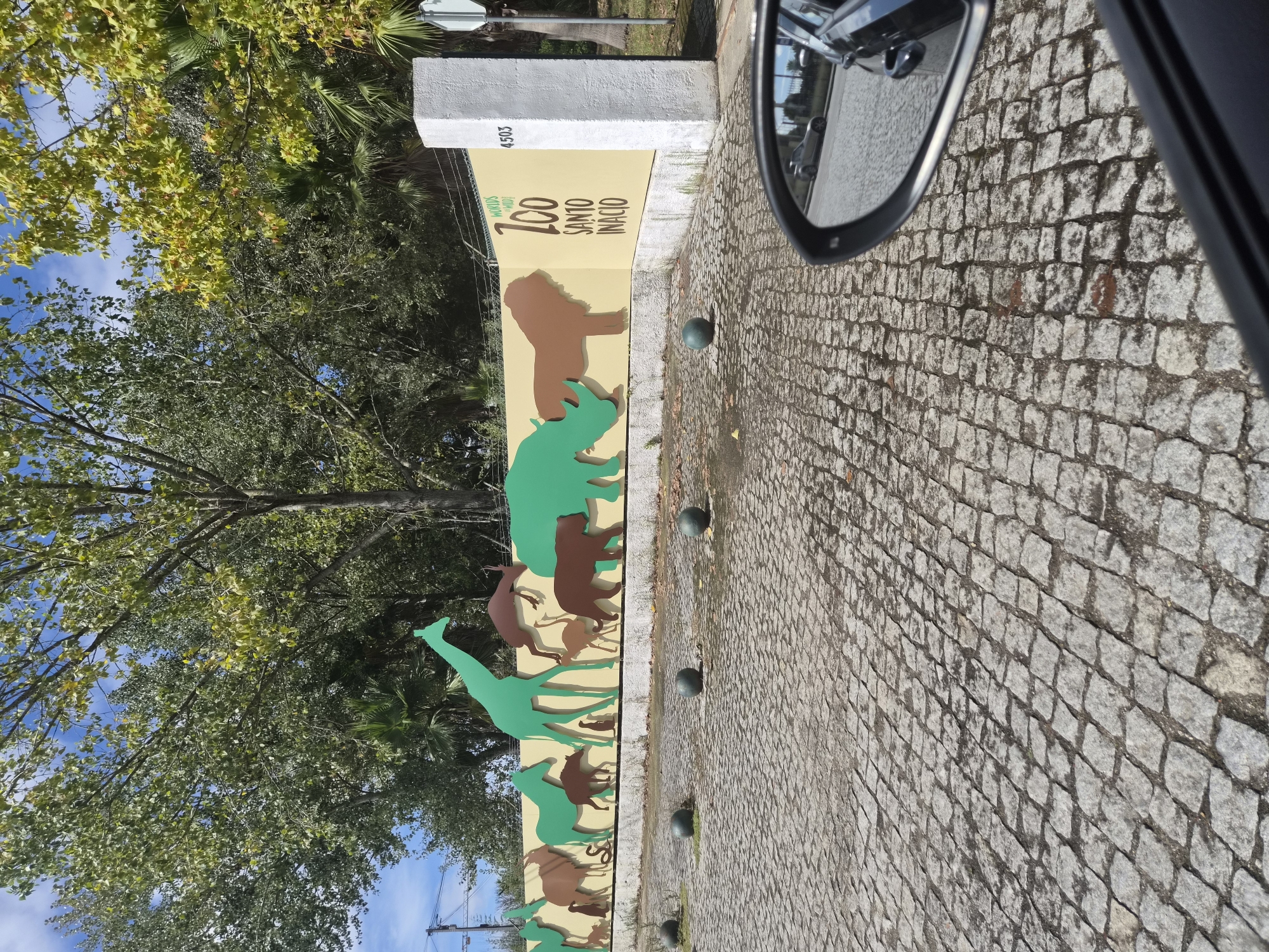 The image shows the entrance to the Zoo Santo Inácio, located in the city of Vila Nova de Gaia, Portugal. A decorative wall features colorful silhouettes of various animals, including a giraffe, rhino, lion, and bear, against a yellow background. The zoo's name is prominently displayed in large text. In the foreground, a cobblestone pavement leads towards the entrance, with several dark, spherical bollards lining the pathway. A white pillar stands to the right of the wall, bearing the number "4503". The scene is framed by the reflection in a car's side mirror, which shows another car and a street in the background, suggesting the photo was taken from inside a vehicle. Lush green trees with dappled sunlight form the backdrop.
