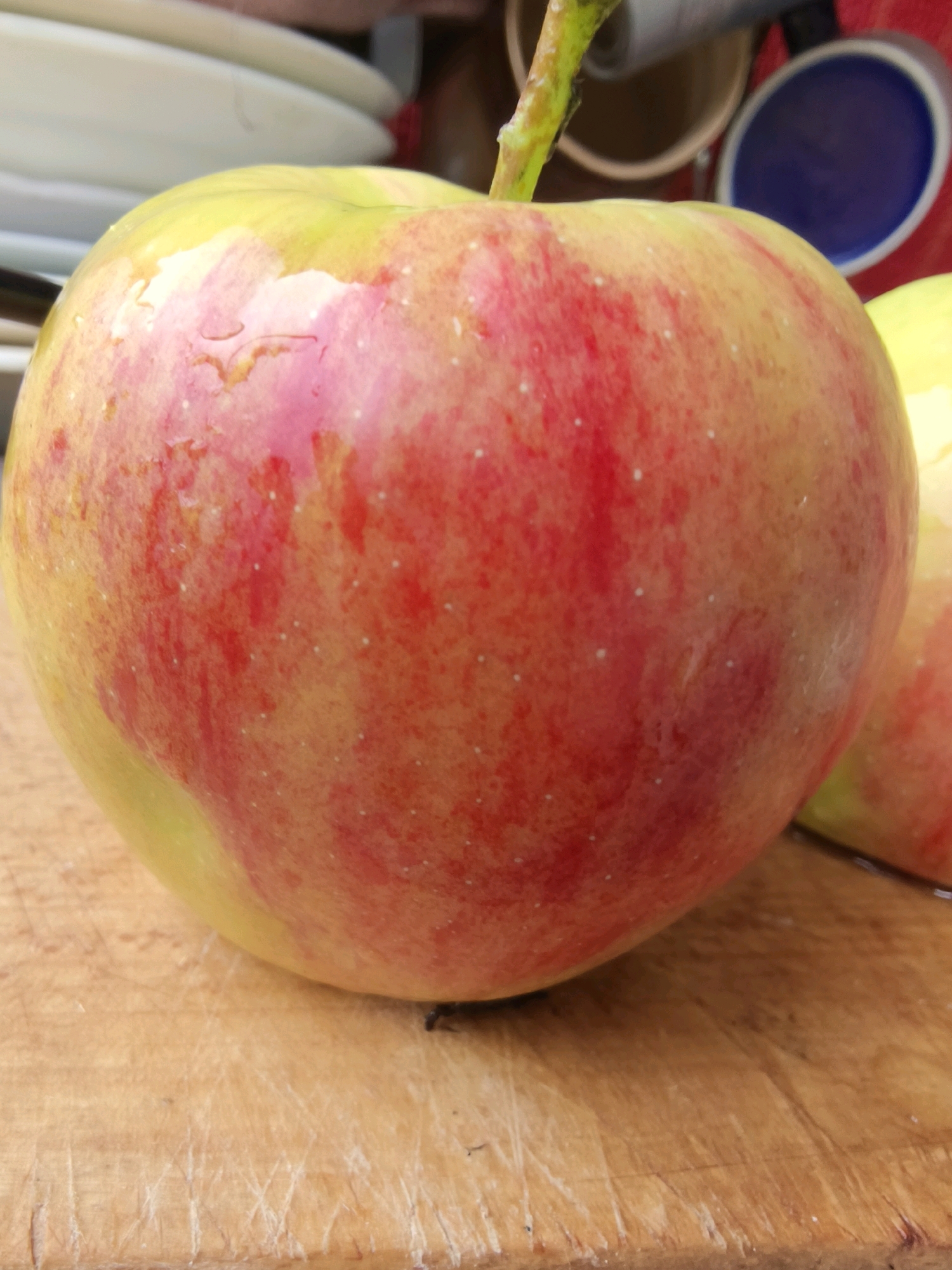 An overhead close-up shot shows a single red and yellow apple with a stem. The apple is on a wooden surface that appears to be a cutting board. In the background, several white plates are stacked, and a blue cup is visible. The lighting suggests an indoor setting. There are no people visible in the image. The image does not contain any elements that allow for the determination of its location within the city of Kryvyi Rih, Ukraine.
