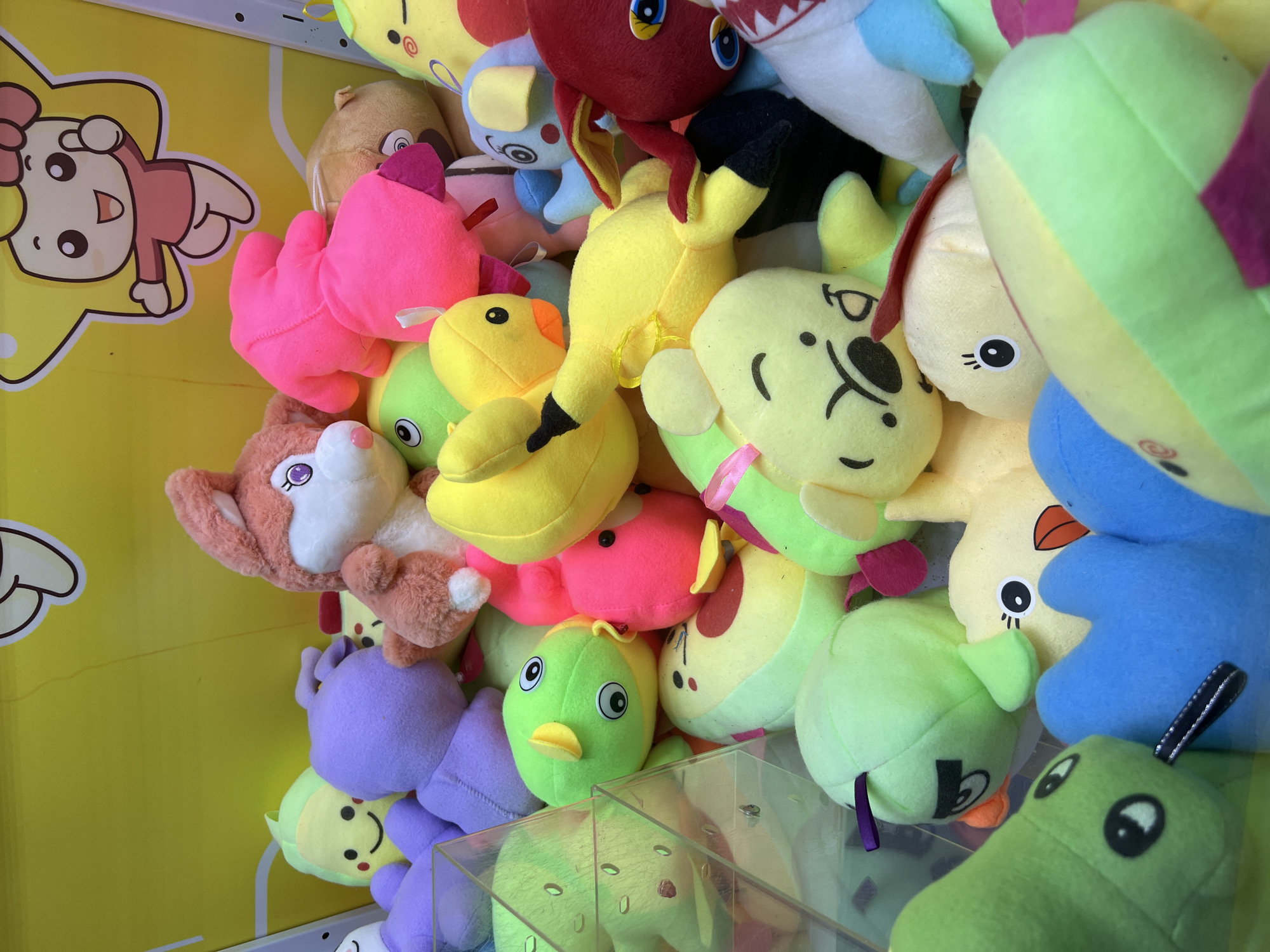A collection of colorful plush toys is densely packed inside a claw machine. The toys are varied in shape and character, including cartoonish animals like a pink rabbit, yellow ducks, and a green bird with angry eyes. There are also abstract shapes like purple and pink blobs, and yellow banana-like figures. Some toys have visible facial features such as eyes, beaks, and smiles. The background of the claw machine is a bright yellow wall with a cartoon drawing of a character in a star. The toys are mostly soft and plush, with bright, saturated colors. This image does not depict any specific location in Eretria Municipal Unit, Greece, as it is an indoor setting of a coin-operated amusement machine.