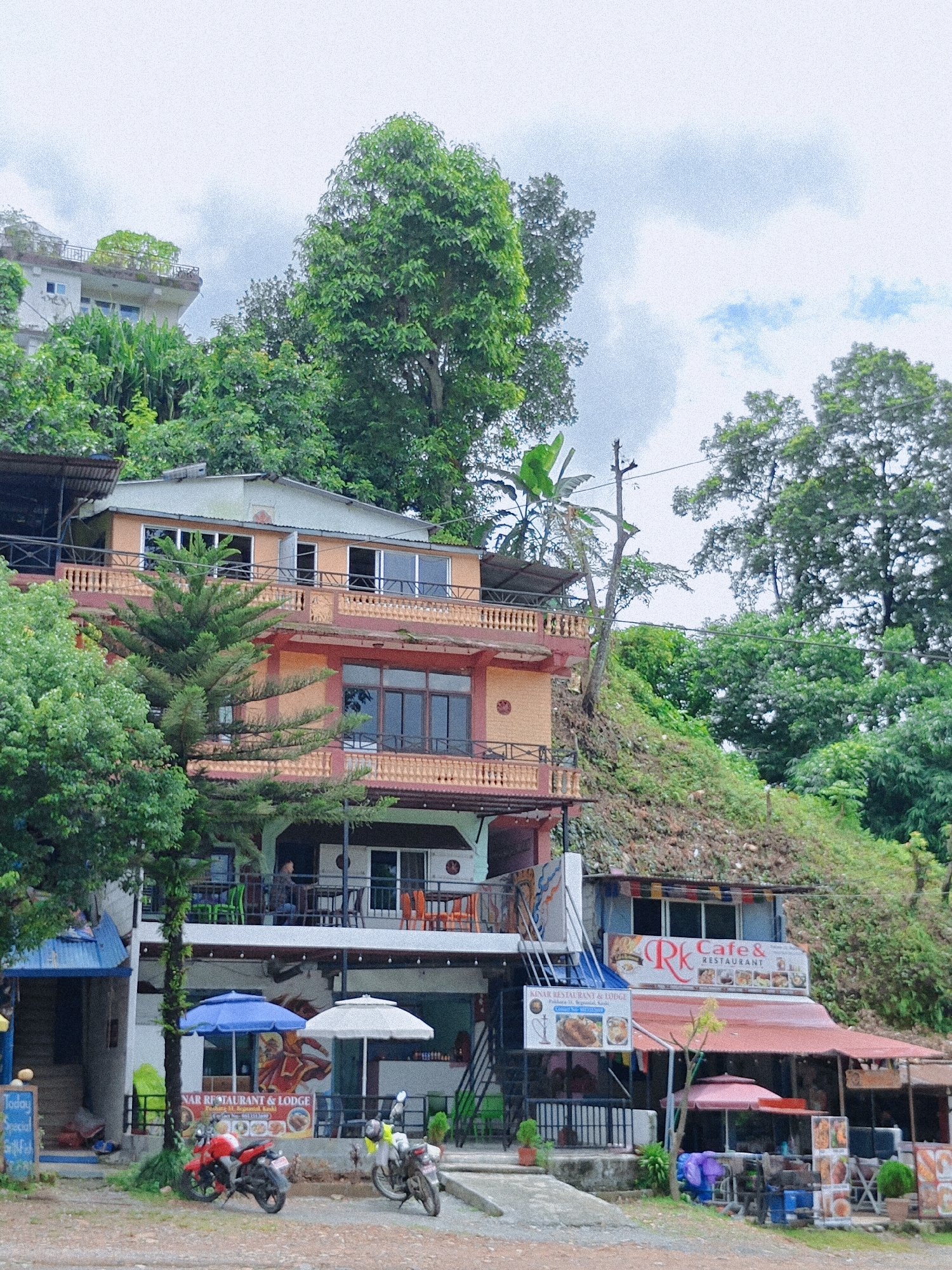 The image depicts a multi-story building complex built into a hillside in Pokhara, Nepal. The buildings appear to house restaurants and lodges, as indicated by signage such as "RK Cafe & Restaurant" and "NAR Restaurant & Lodge." Several levels of balconies and patios are visible, some with outdoor seating. Trees and lush vegetation surround the structures, with the hillside itself covered in greenery and dirt. In the foreground, two motorcycles are parked, a red one and a silver one. Various umbrellas are set up outside one of the establishments. The sky is overcast with some patches of blue. A few people are visible in the background, seated at tables on a balcony. The overall impression is of a commercial establishment situated in a hilly, tropical or subtropical environment.