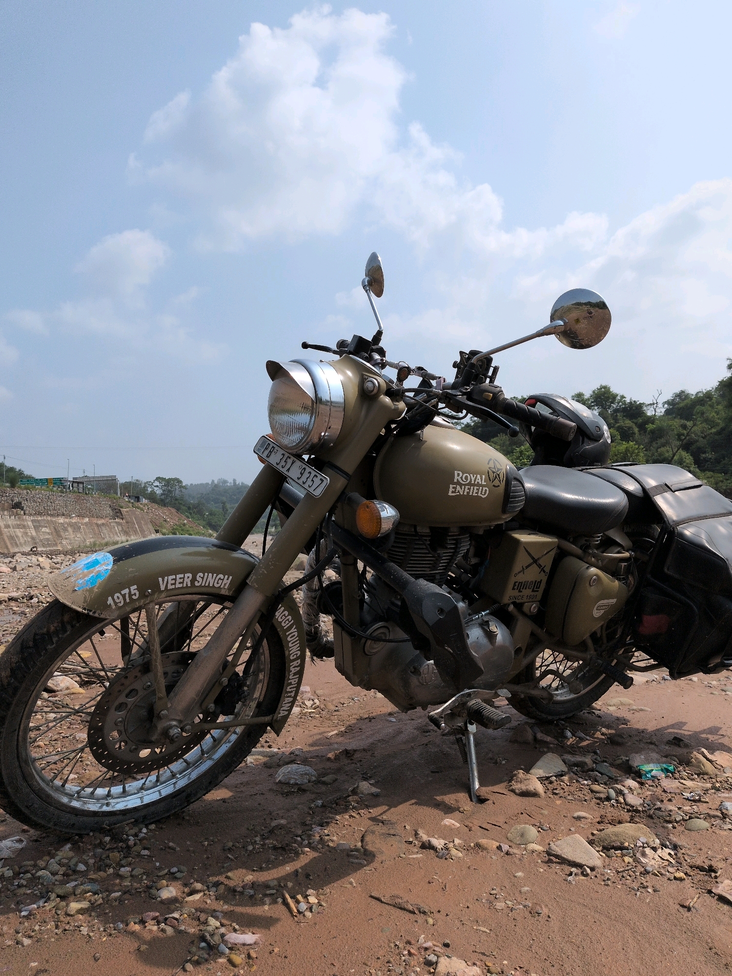 A khaki-colored Royal Enfield motorcycle is parked on a dirt and rocky ground. The motorcycle has "ROYAL ENFIELD" branding on its fuel tank and a "CLASSIC" decal on its side. The front fender has "VEER SINGH" and "1975" written on it, along with some blue markings. The license plate reads "PB 35Y 9357". A black helmet rests on the seat. In the background, there is a concrete wall, trees, and a bright sky with scattered clouds. The location is identified as Jaanta, India.