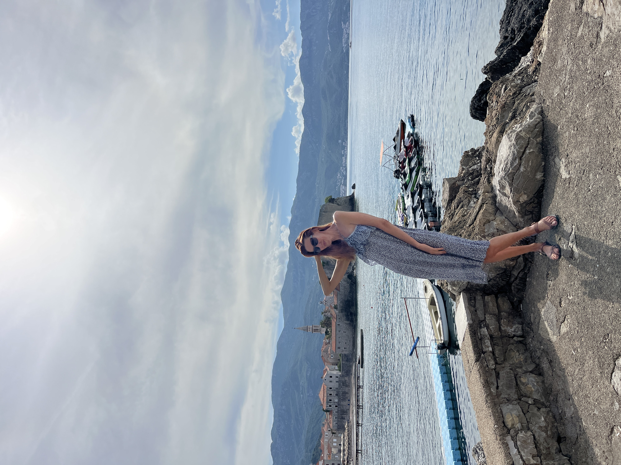 A scenic photograph captures a woman posing on a bright, sunny day in Budva, Montenegro. The image, horizontally oriented, features a woman with reddish-brown hair, wearing sunglasses, a patterned sleeveless dress, and sandals. She is positioned on the right side of the frame, standing on a concrete and rocky embankment, with one hand casually raised towards her head as she looks towards the camera with a relaxed expression.
To her left, the calm, blue waters of the sea extend, dotted with several small boats and jet skis moored near the shore, and a white boat tied to a blue floating dock. Across the water in the mid-ground, the historic old town of Budva is visible, characterized by traditional stone buildings with red-tiled roofs and a prominent bell tower or steeple. Towering mountains, partially obscured by scattered white clouds, form a majestic backdrop to the coastal settlement.
The sky is bright and mostly clear, indicating fair weather. The scene conveys a tranquil and picturesque atmosphere, typical of a leisurely moment by the Adriatic coast. There is no visible text in the image.