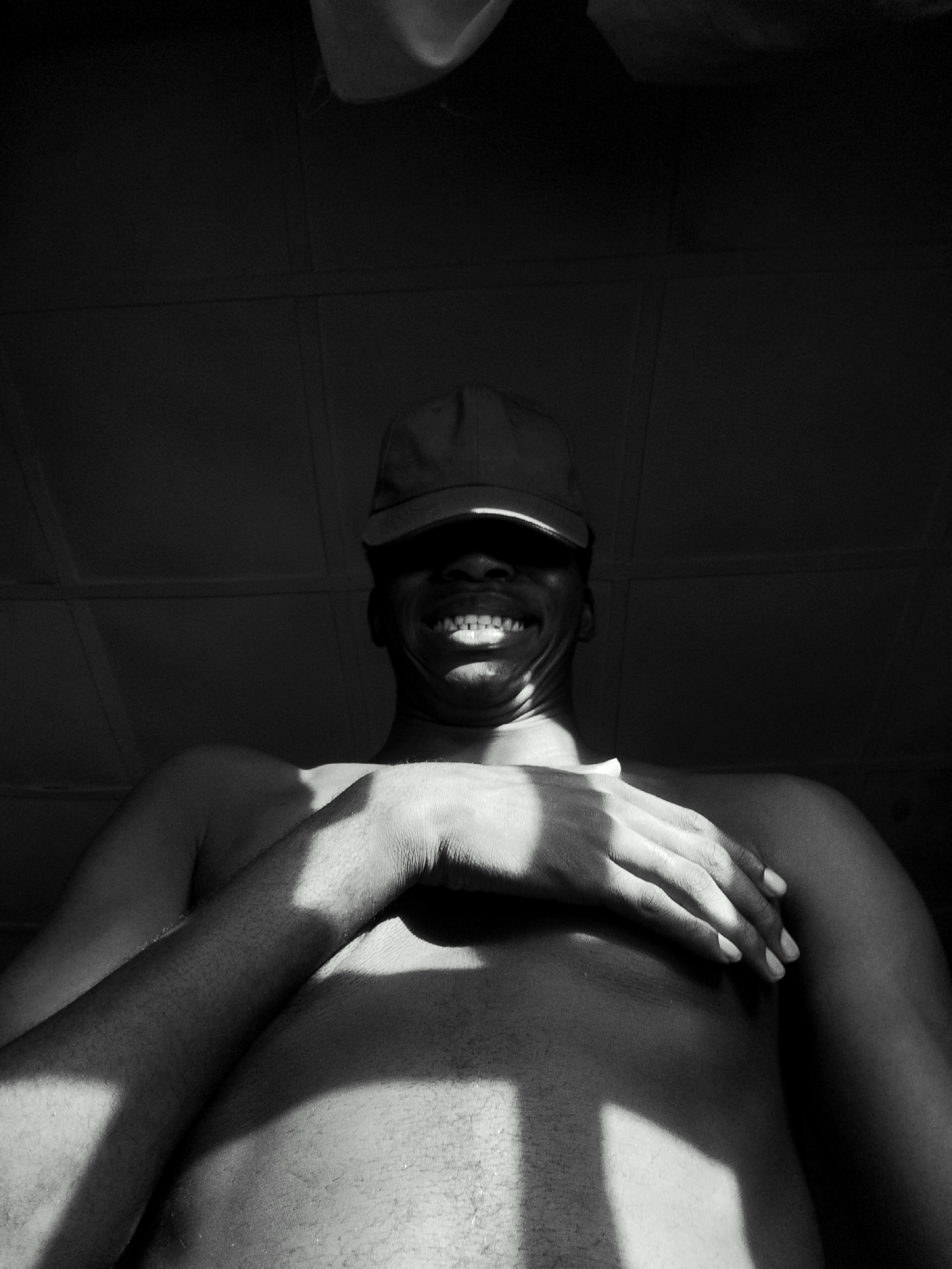 A black and white, low-angle shot depicts a dark-skinned, shirtless person wearing a baseball cap, their eyes obscured by the cap's brim. The person is smiling, showing their teeth. Their right hand is placed on their left shoulder, with a strong light casting shadows across their body. The background is a dark, possibly tiled ceiling. There is no available information indicating the location of the scene is in Keffi, Nigeria.