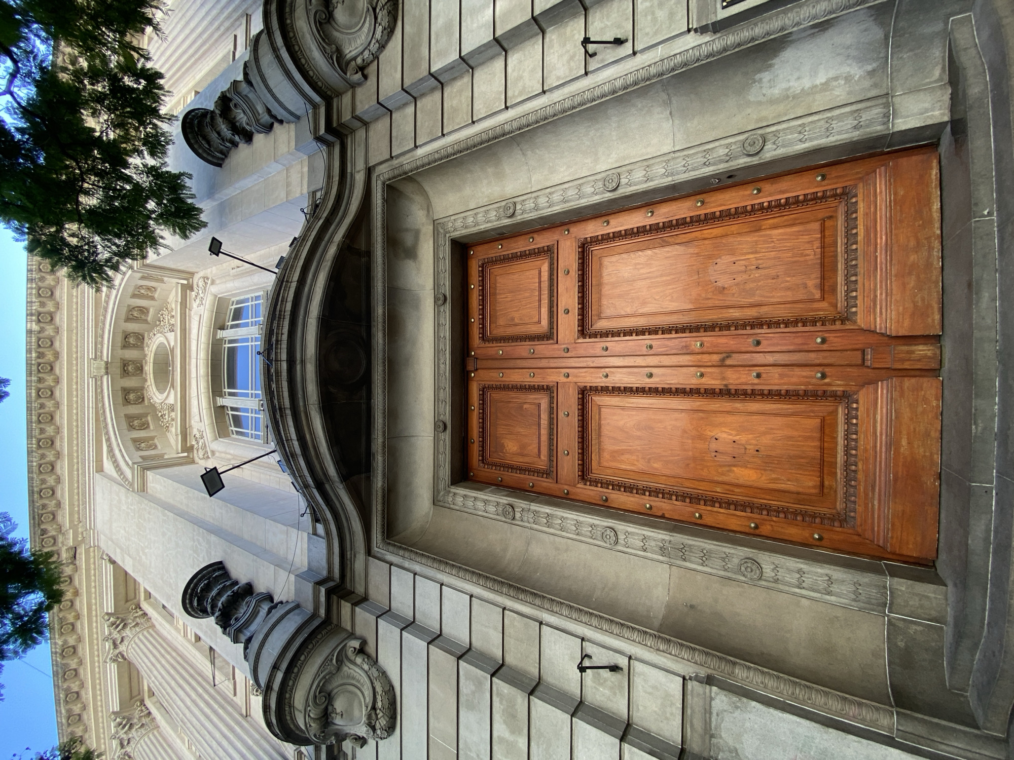 The image shows an ornate wooden door, likely the entrance to a building in Buenos Aires, Argentina. The door is made of two large wooden panels with raised rectangular details, bordered by decorative carvings and studded with brass rivets. The doorframe is constructed of light-colored stone, adorned with geometric patterns and circular motifs. The building façade features intricate architectural elements such as fluted columns, decorative moldings, and sculpted details. A portion of a tree is visible on the left side of the frame. Above the door and facade are windows, with bright blue visible from the sky. Additionally, there is a black camera or security light affixed to the wall of the building.