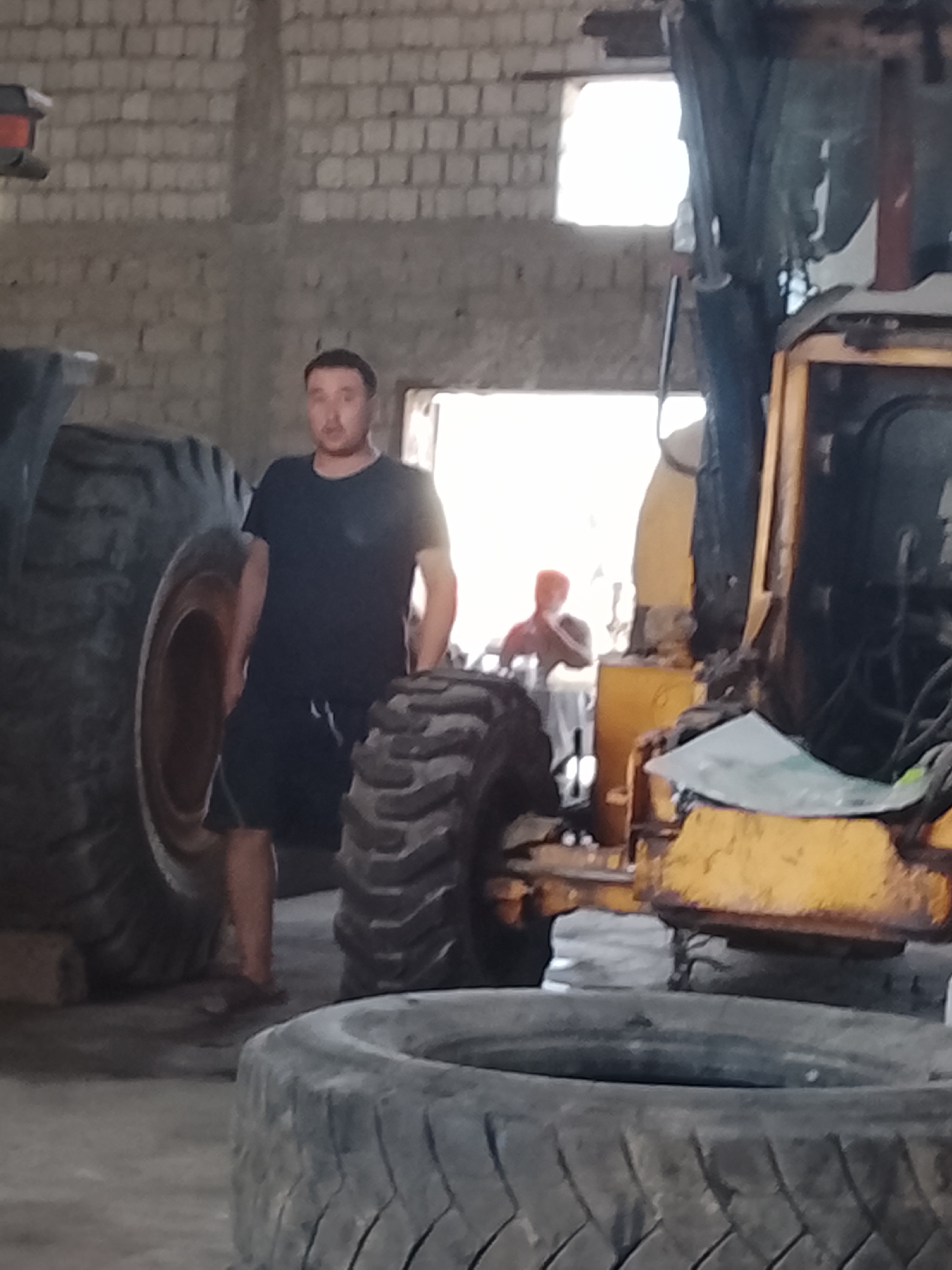 A man in a black t-shirt and shorts stands in what appears to be a garage or workshop in Sabir Rakhimov, Uzbekistan. He is positioned between the large tire of a heavy machine, possibly a tractor or loader, and another large tire in the foreground. In the background, through an opening, another person wearing an orange head covering is visible, sitting and looking towards the viewer. The environment is industrial, with exposed brick walls and natural light coming through the opening. The heavy machinery and tires suggest a setting related to vehicle repair or maintenance.