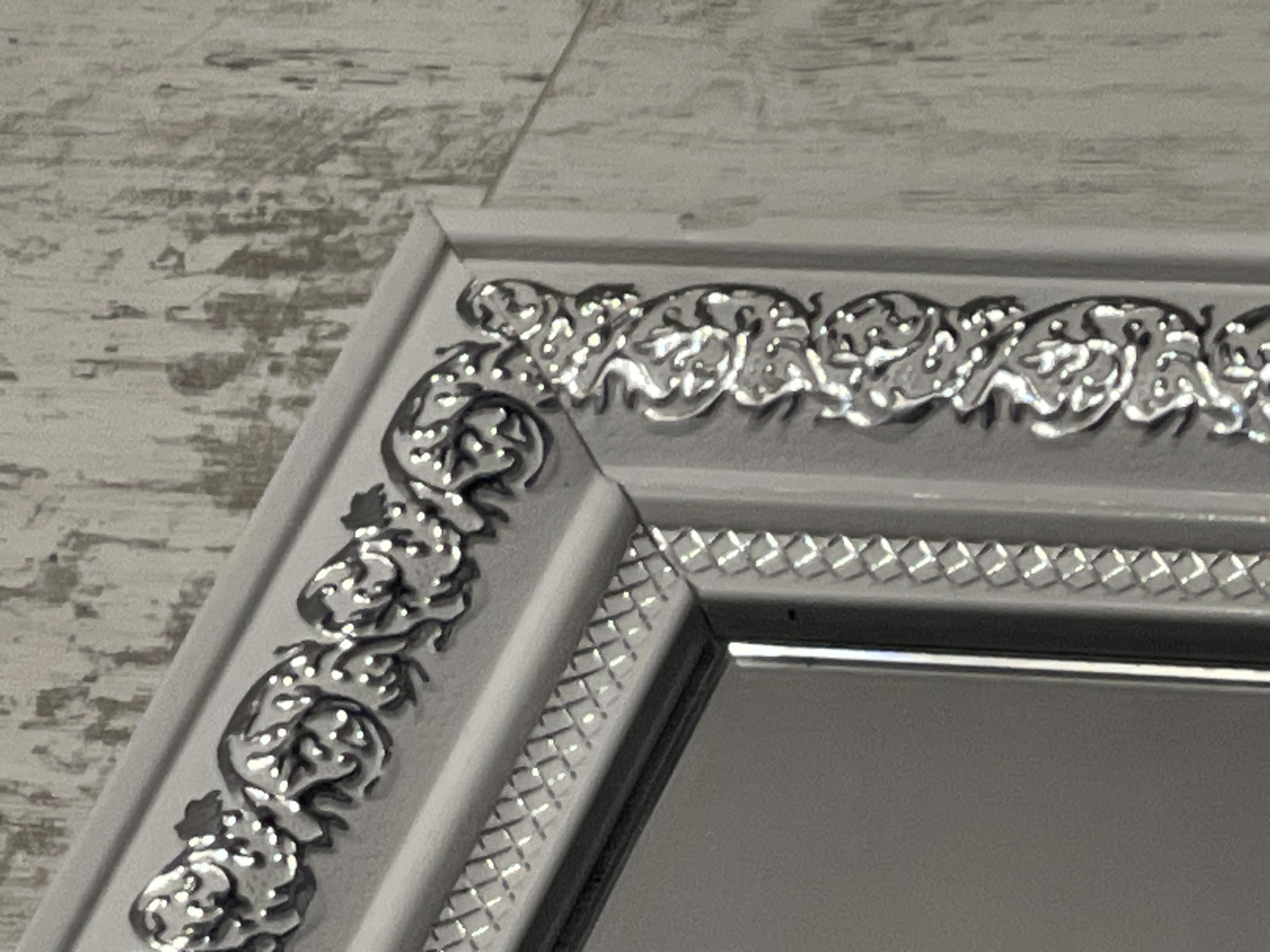 A close-up shot of a mirror frame shows ornate, silver decorative trim with swirling patterns on a light gray frame. The frame is positioned in front of a textured, gray wall. The mirror reflects a muted gray surface.