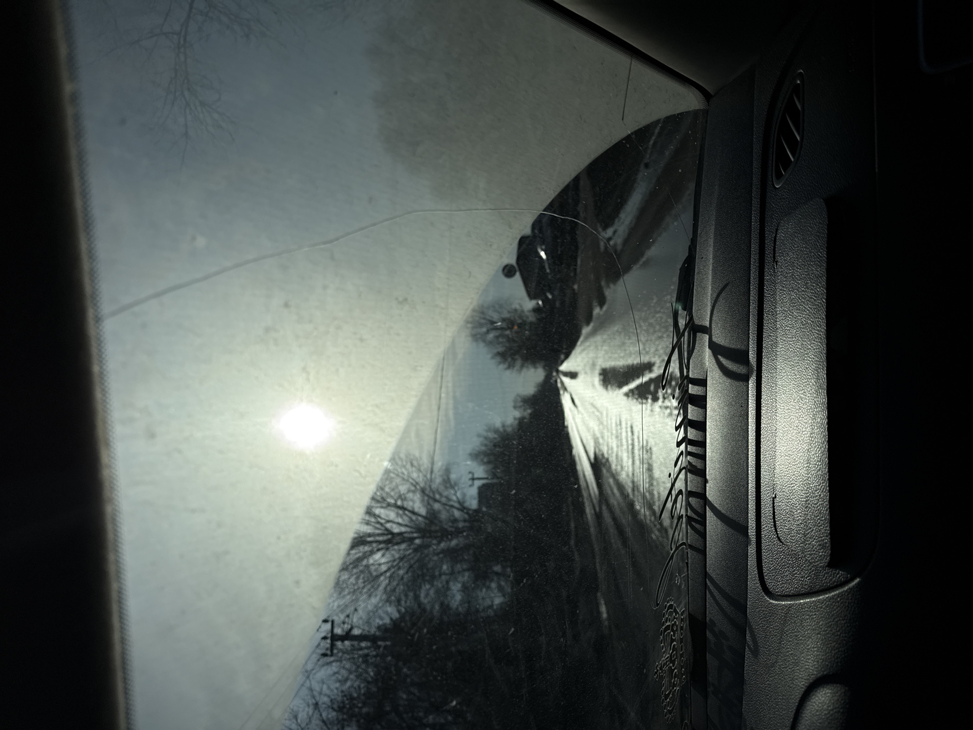 The image, taken from inside a vehicle, shows a view through a cracked windshield. The scene outside depicts a road in Chaikovsky, Russia. It appears to be a wintery day with bare trees visible along the roadside. In the foreground, part of the vehicle's interior, including the dashboard and an air vent, are visible. A silhouette of trees and utility poles is reflected on the windshield. The sun is shining through the windshield, creating a bright spot in the center.