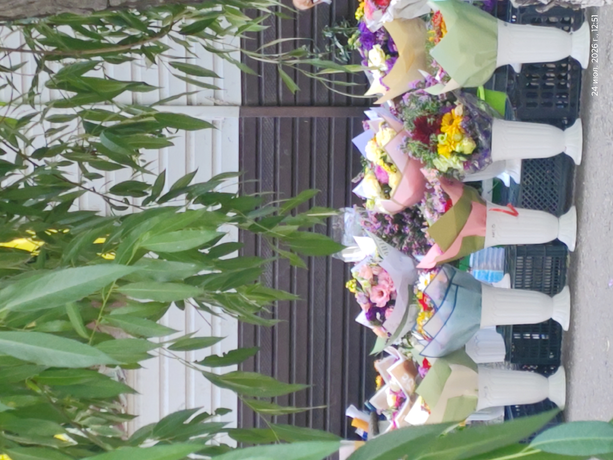 A street flower stall in Kokshetau, Kazakhstan, is depicted, featuring an assortment of floral bouquets. The stall is set up outdoors, with a wooden fence in the background. The bouquets are arranged in various decorative wrappers and are placed in white vases. Some of the flowers are vibrant, with colors like pink, yellow, purple, and red. The scene appears to be captured on a sunny day, with sunlight filtering through the green foliage of a tree that hangs over the stall. The image is dated "24 июл. 2026 г., 12:51".