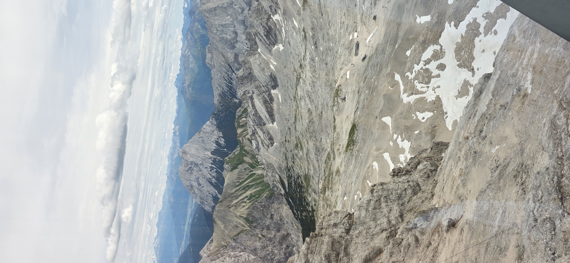 This is a high-angle, vertical, full shot of a vast mountainous landscape, likely taken from an elevated vantage point such as a cable car or viewpoint. The scene is dominated by rugged, grey rock formations and sparse vegetation, with patches of snow scattered across the terrain.
In the foreground, a steep, rocky cliff face occupies the left side of the frame, with visible weathering and loose scree. The midground opens up to a wide, undulating valley floor composed of gravelly soil and rock debris. Small remnants of snow are interspersed across this area, forming irregular white patches. There are a few scattered buildings and structures visible in the midground, some appearing to be ski lifts or facilities, and a narrow, paved path or road winds through the valley.
The background reveals a layered panorama of distant mountain ranges, fading into a hazy blue due to atmospheric perspective. The highest peaks in the far distance are capped with snow and appear sharp against the sky.
The sky is overcast with a thick layer of grey clouds, interspersed with lighter, wispy clouds. The light is diffused, suggesting either a cloudy day or early morning/late afternoon.
While the image depicts a natural mountain environment, its location in Grainau, Germany, implies it is likely a view of the Zugspitze massif, Germany's highest peak, which is accessible from Grainau via cable car. The presence of ski lift structures further supports this interpretation of a popular mountain resort area. There are no discernible people or animals in the frame.
