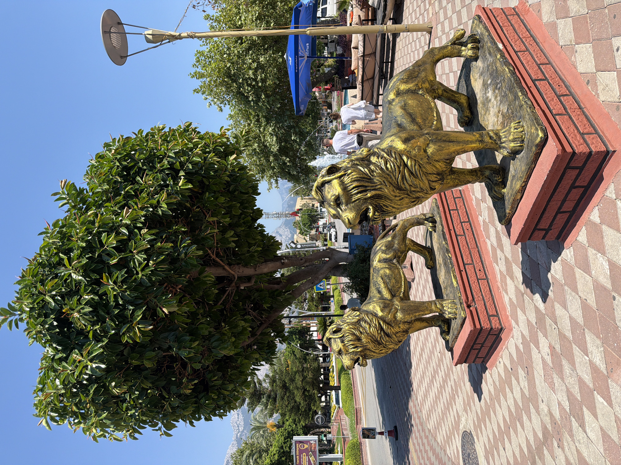 Two golden lion statues stand prominently on a patterned brick walkway in Kemer, Turkey. The statues are placed on red brick pedestals, and both are facing to the left of the frame. A large, leafy green tree dominates the center of the image, casting shadows on the walkway. In the background, a bright blue umbrella shades an outdoor seating area with several tables and chairs. A couple, a man and a woman, walk away from the camera, towards a fountain visible behind them. The scene is set against a clear blue sky, with mountains visible in the distance. Other elements include a street lamp, a street sign, and parked cars.