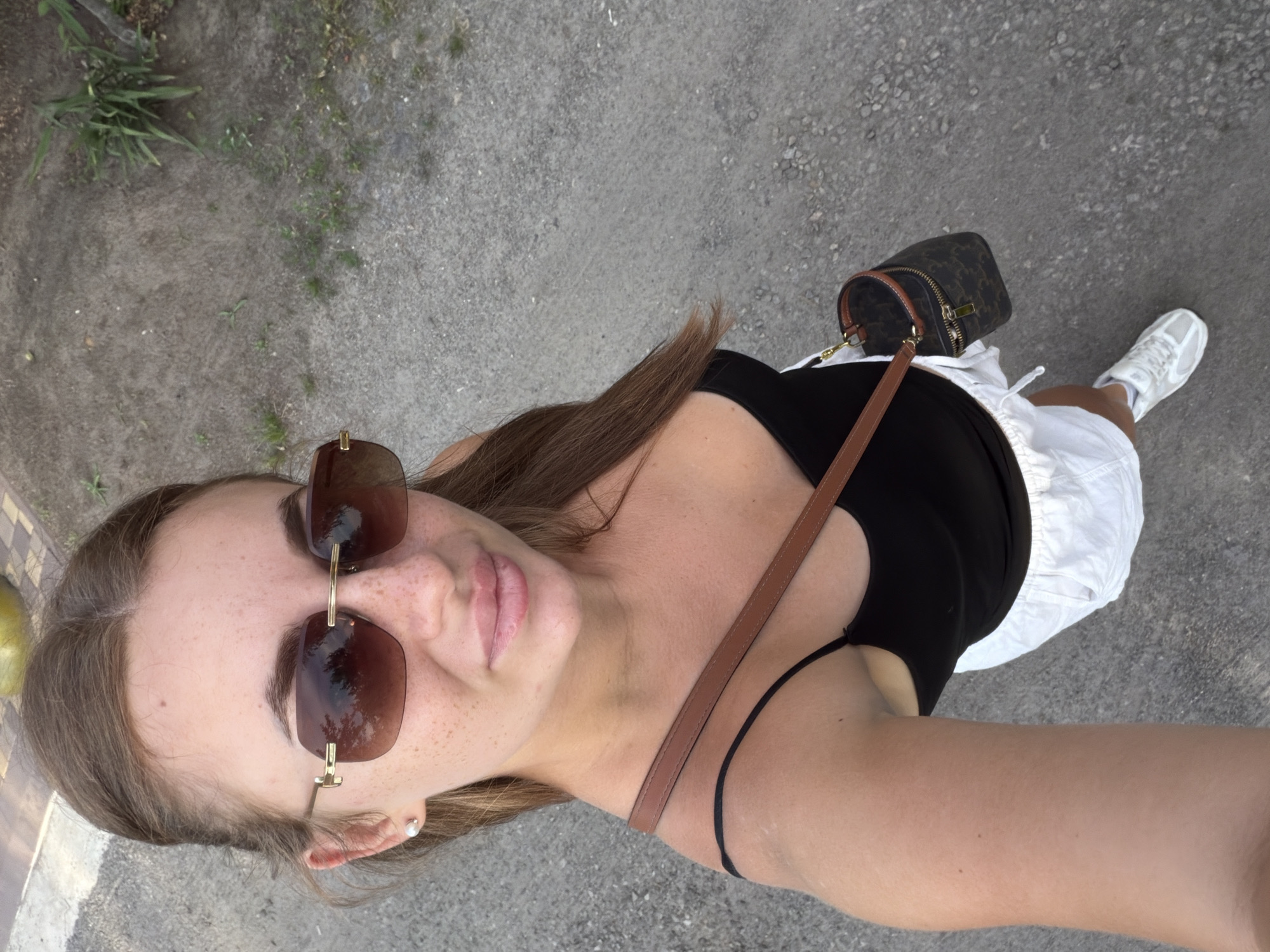 A woman with brown hair and freckles is taking a selfie outdoors. She is wearing sunglasses, a black tank top, white shorts, and white sneakers. She has a small brown bag with a strap across her chest. The ground is covered in gravel and small plants. The lighting suggests it is daytime. It is not possible to determine the specific location within Dnipro, Ukraine, from the visual information provided.