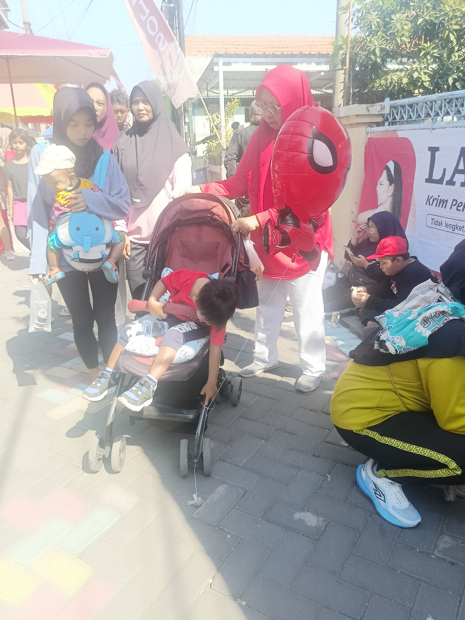 The image depicts an outdoor scene in Gedangan, Indonesia, on a paved walkway with a patterned brick surface. The lighting suggests it is daytime, with a bright sky and noticeable shadows. In the foreground, a child wearing a red t-shirt and dark shorts is seated in a stroller. The stroller is a dark, reddish-brown color. To the left of the stroller, a woman wearing a light blue long-sleeved shirt and dark leggings carries a baby in a blue and gray baby carrier. The baby is wearing a light-colored hat and yellow clothing. Behind the woman, another woman in a black hijab and pink top looks forward. Further back, more people are visible, including a woman in a red hijab holding a Spiderman balloon. To the right of the frame, a person in a yellow top and black pants with a yellow stripe is crouched down, looking at a mobile phone. A banner with Indonesian text and an image of a woman is visible in the background. The environment appears to be an urban or semi-urban area, possibly a market or event, given the presence of tents and stalls in the background.