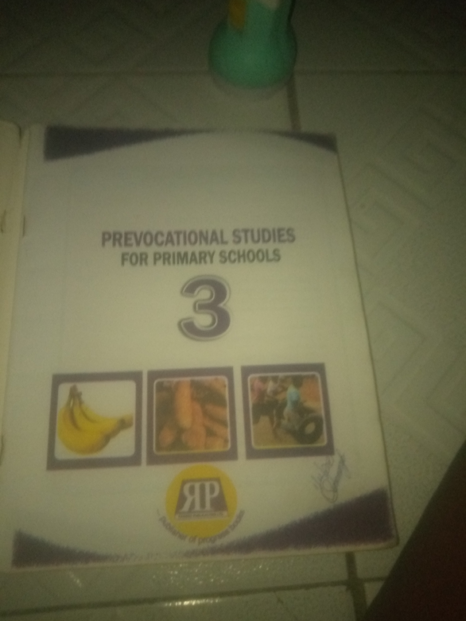 The image shows the front cover of a textbook titled "PREVOCATIONAL STUDIES FOR PRIMARY SCHOOLS 3". The cover features a large number "3" in the center, with smaller images of bananas, carrots, and a person riding a motorcycle in the lower section. Below these images, there is a circular logo with the letters "RP" inside, and text that appears to read "Publisher of Progress Books". The textbook is placed on a tiled surface, possibly a floor or table. The lighting is dim, and the image is slightly out of focus, suggesting it was taken indoors. There is no identifiable information within the image that would place it specifically in Jauro Yinu, Nigeria.