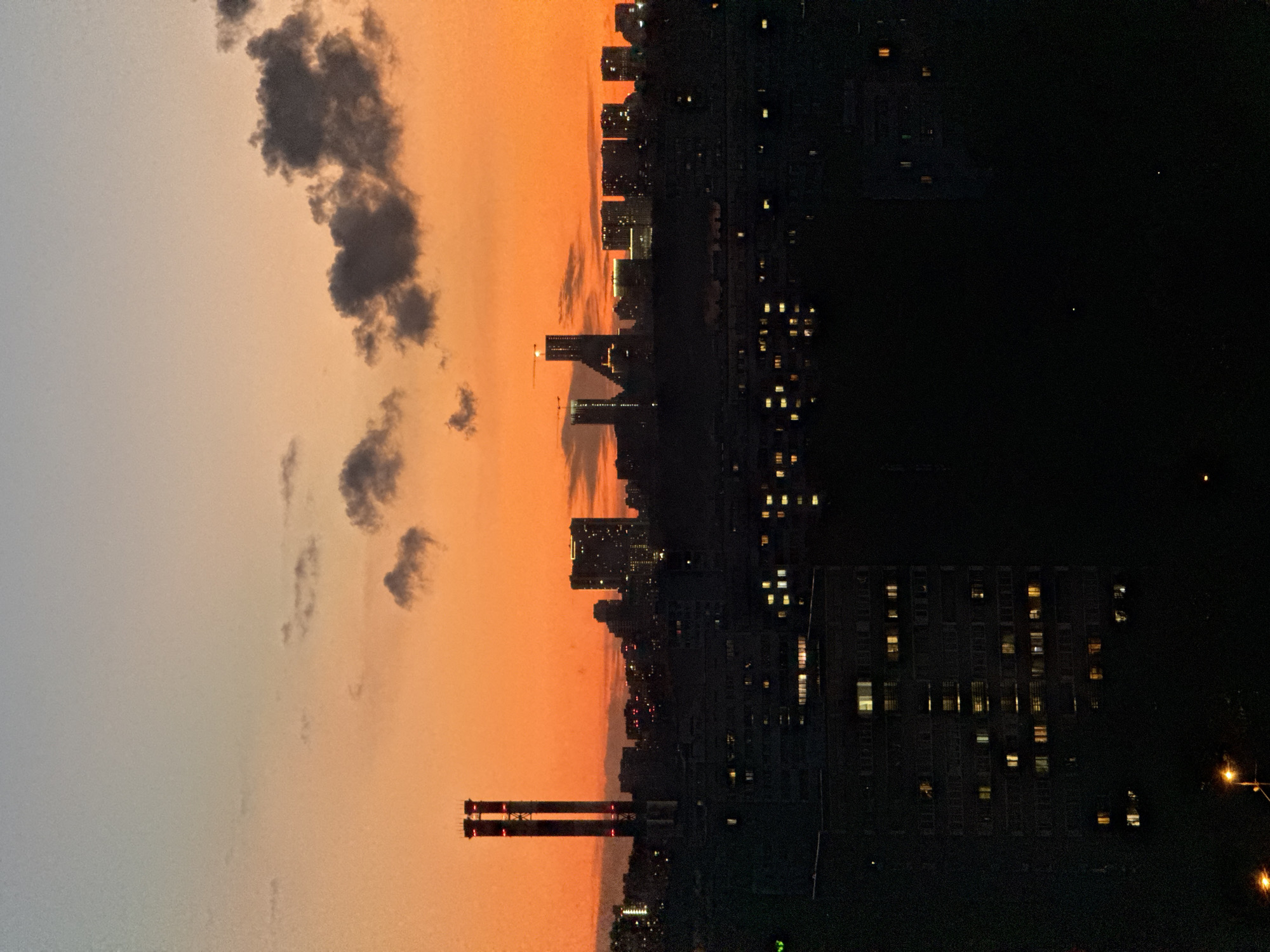 The image captures a city skyline at sunset, likely Moscow, Russia, given the architectural styles and the presence of tall, distinct structures. The sky transitions from a pale blue at the top to a vibrant orange near the horizon, illuminated by the setting sun. Silhouetted against this colorful sky are various buildings, including a prominent pair of tall industrial chimneys with red lights near their tops, and a cluster of high-rise buildings with illuminated windows in the distance. In the foreground, the dark silhouettes of lower-rise apartment buildings are visible, some with windows lit up, suggesting habitation. The overall scene depicts an urban environment at dusk, with the transition from daylight to night. No people or specific actions are discernible in the image.