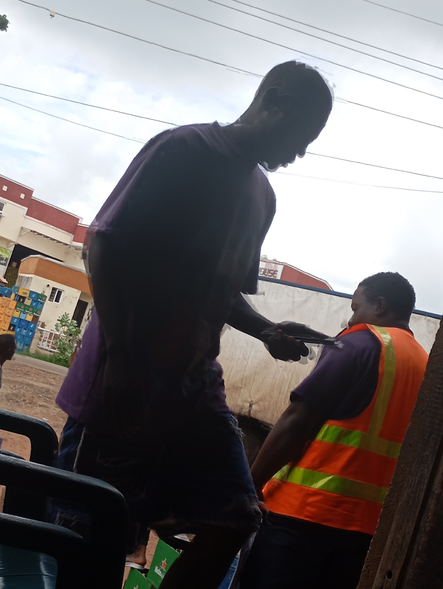 In this close-up, low-angle shot taken outdoors, two individuals are prominently featured, silhouetted against an overcast sky. The person in the foreground, to the left, is partially obscured by their dark purple t-shirt and blue denim shorts. Their head is turned downward, and their right arm is extended, holding a smartphone. To the right, the second individual wears a bright orange safety vest with reflective yellow stripes over a purple shirt. Their body is angled away from the camera, and their head is turned to the side, suggesting an interaction between the two. In the background, several buildings are visible, including one with a red and white facade and another that appears to be a warehouse. Stacks of colorful crates are piled near the buildings, and a few green Heineken bottles are visible at the bottom of the frame. The environment appears to be an urban or industrial area, possibly a loading dock or a street with commercial establishments.