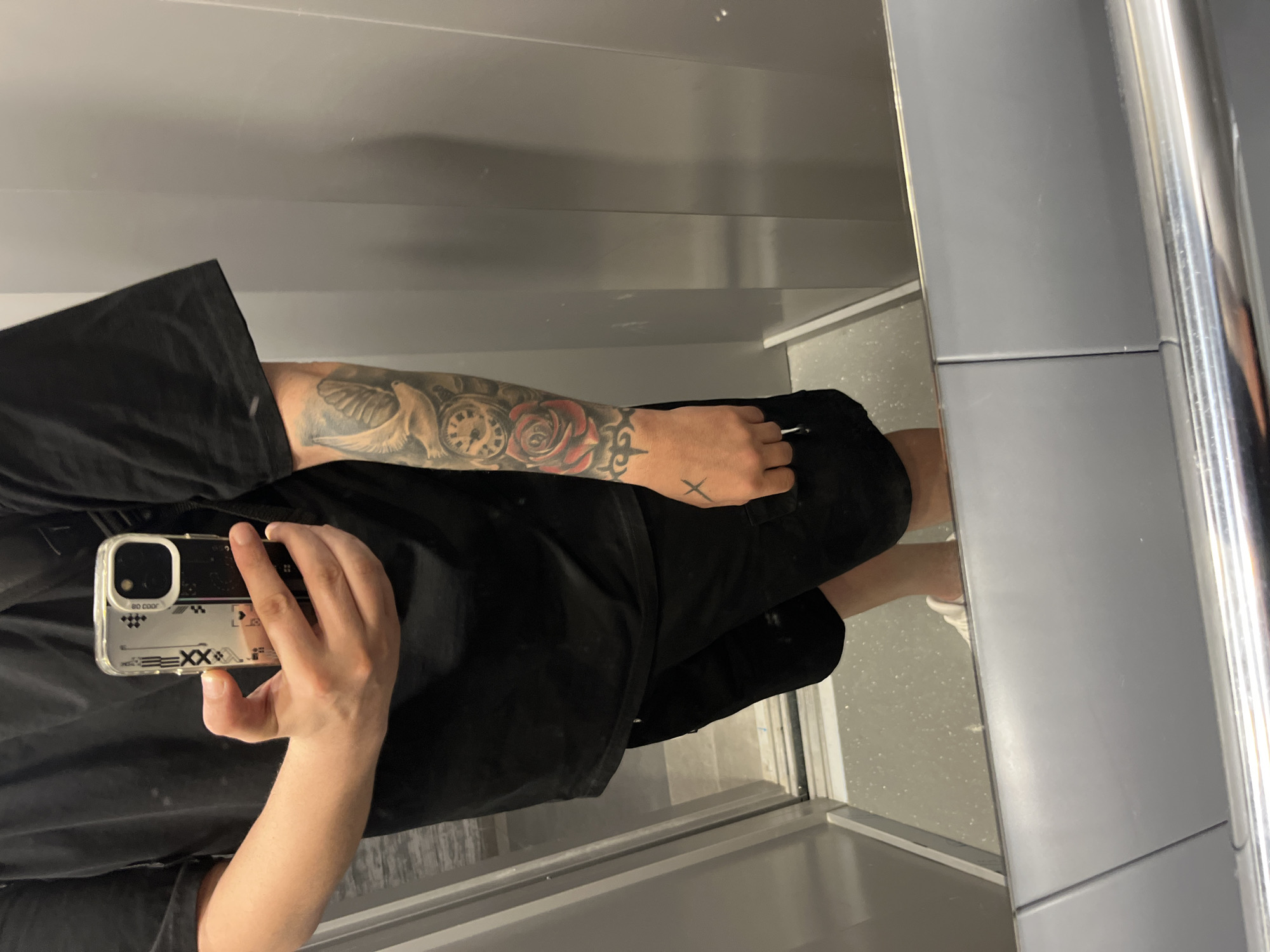 The image displays a mirror selfie of a person inside an elevator located in Varna, Bulgaria. The person is reflected from the chest down, dressed in a plain black short-sleeved t-shirt and black cargo shorts, with white sneakers partially visible in the lower reflection. The right arm (viewer's left) is heavily tattooed, featuring a white dove with spread wings near the shoulder, a circular pocket watch, a prominent red rose with green leaves, and a thorny band design towards the wrist. A black 'X' symbol is tattooed on the back of the right hand. The individual is holding a smartphone, appearing to be an iPhone, horizontally with their left hand (viewer's right). The phone is encased in a clear or translucent cover decorated with a "NBD COLOR" text snippet, a "EXX" logo with double 'X' symbol, a checkerboard pattern, and playing card suit symbols (spades, clubs, diamonds, hearts). The elevator interior features light grey paneled walls with vertical seams, a light grey speckled floor, and a polished metal handrail running along the bottom. Metallic door frames are visible on the left side of the reflection.