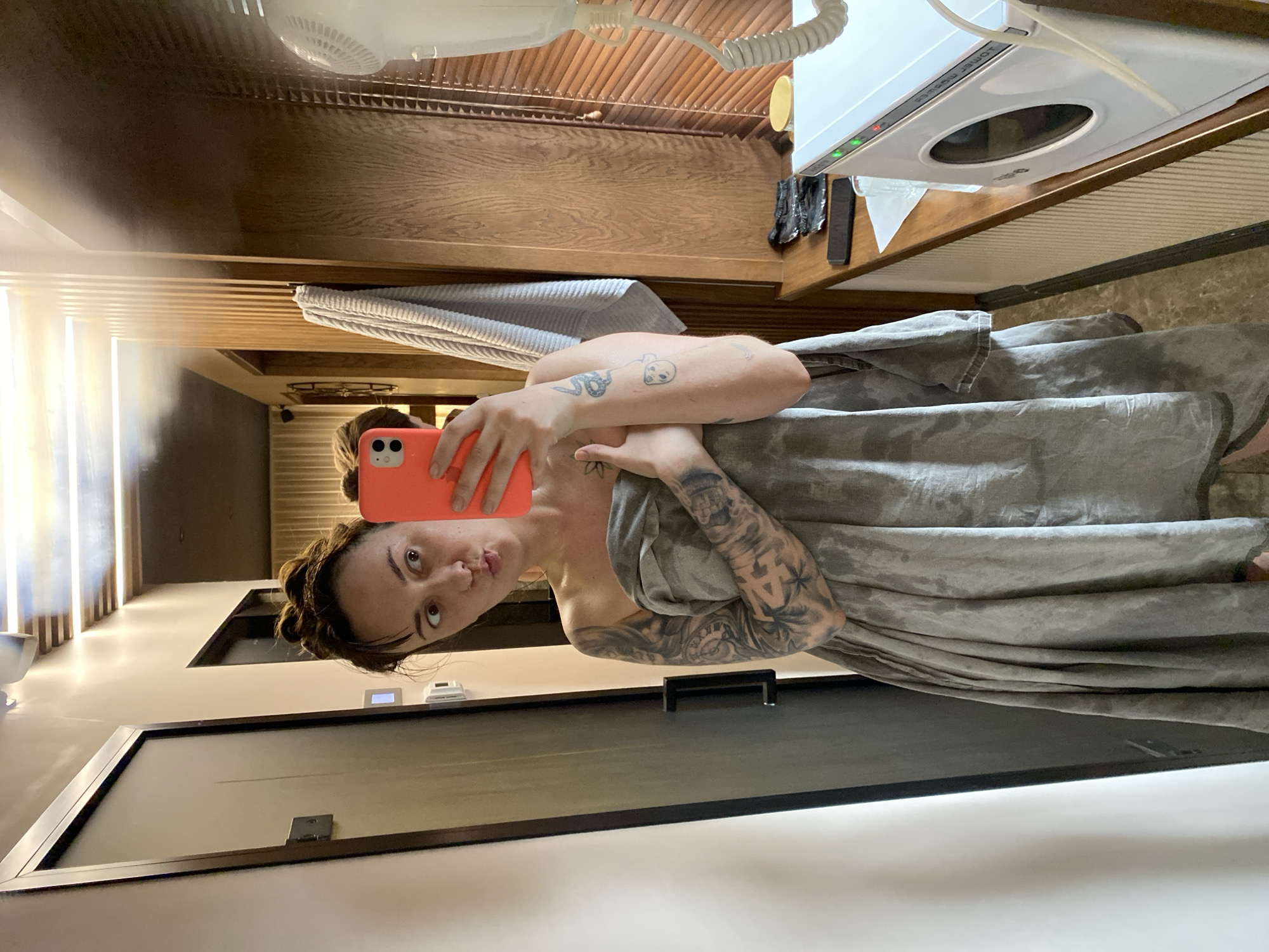 A person stands in a bathroom with the image tilted such that the left side is the top. The person holds a peach-colored smartphone, taking a selfie. The person is wrapped in a gray towel or sheet. Tattoos are visible on the arms.
The bathroom contains a black-framed glass shower door. Beyond the door, a light wall with vertical lighting is visible. The wooden ceiling has a hairdryer mounted on it. A white dryer is on a wooden shelf along the upper right side. A white towel hangs near the person, and a gray towel rests on the wooden ceiling.