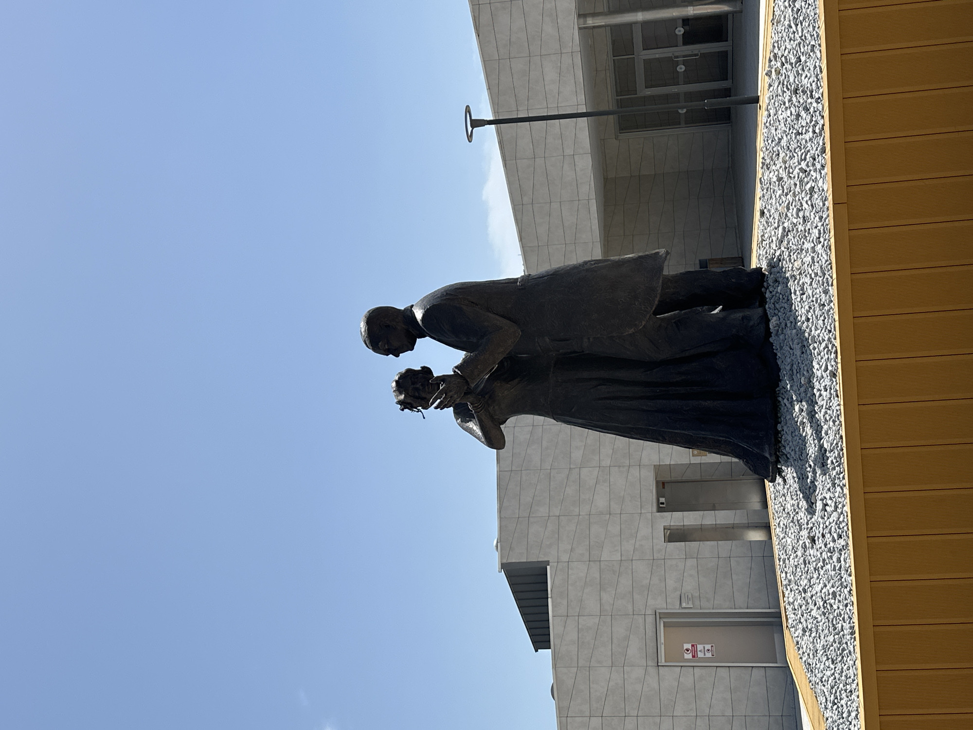 The image shows a bronze sculpture of a man bending down to give flowers to a woman, standing against a backdrop of a modern building facade and a clear blue sky. The sculpture is placed on a bed of small gray stones bordering a wooden-paneled section of the building. The building has a light gray facade with a geometric pattern. A streetlight extends from the building above the sculpture. A glass window is visible on the building, and a door with signage can be seen below it. The sculpture is most likely located in Muğla, Turkey.