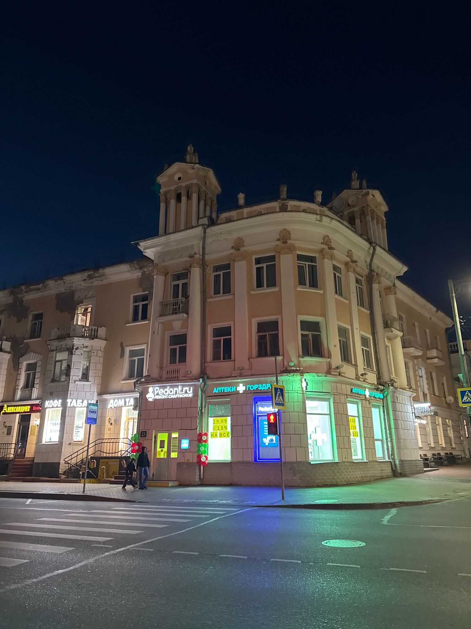 The image shows a nighttime view of a building on a city street in Tver, Russia. The building is a multi-story structure with classical architectural elements, including columns and decorative carvings. Several businesses are visible on the ground floor, indicated by illuminated signs. These include "АПТЕКИ ГОРЗДРАВ" (Apteki Gorzdrav - pharmacies), "Pedont.ru", and other shops with Cyrillic lettering such as "КОФЕ ТАБАКА" (Kofe Tabaka - coffee and tobacco) and "ДОМ ТА" (Dom Ta). A pedestrian crossing is prominent in the foreground, with streetlights and building lights illuminating the scene. A traffic light showing a red pedestrian signal is visible on a pole. Two individuals are walking across the street near the building entrance. The sky is dark, indicating it is night.