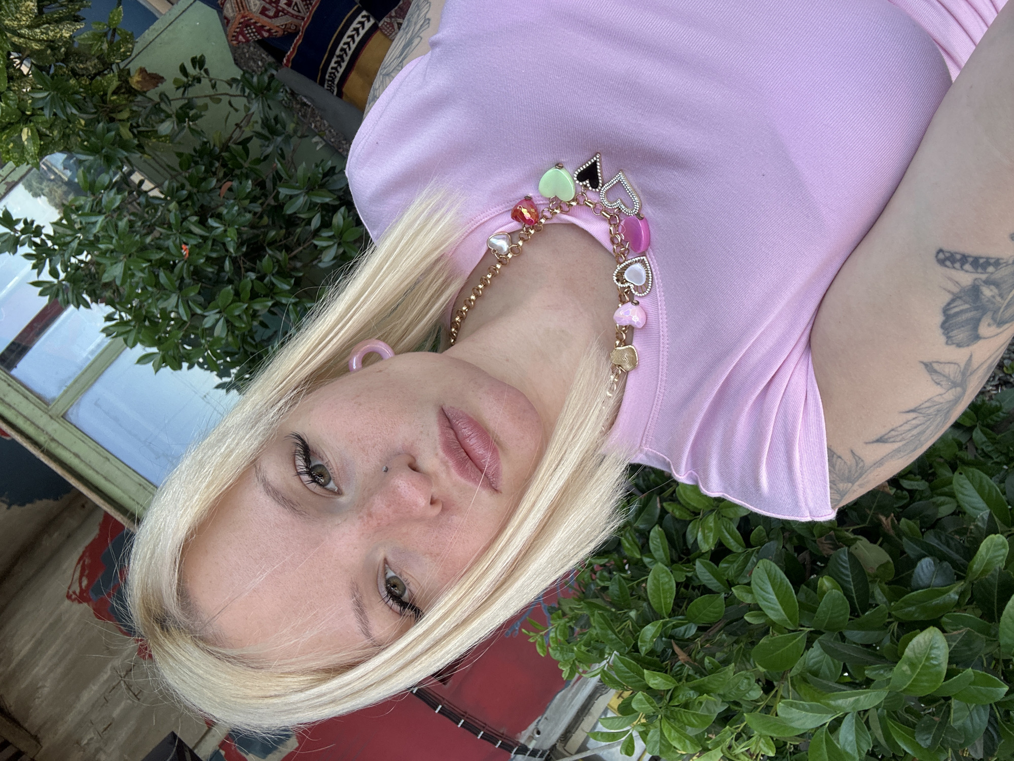 A young woman with blonde hair and light skin, wearing a pink t-shirt and a heart-shaped charm necklace, poses for a selfie outdoors. She has makeup on and a piercing on her nose. She has tattoos on her left arm. Behind her, there are green bushes and a building with red and green elements. The image appears to be taken in Belgrade, Serbia, likely in a garden or outdoor seating area of a cafe or restaurant.
